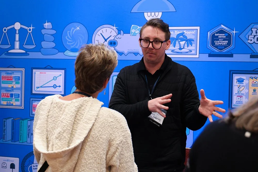A man with glasses and a black jacket is speaking to a woman with short hair, wearing a cream-colored sweater, at a conference or event with a blue backdrop featuring graphs, icons, and awards.