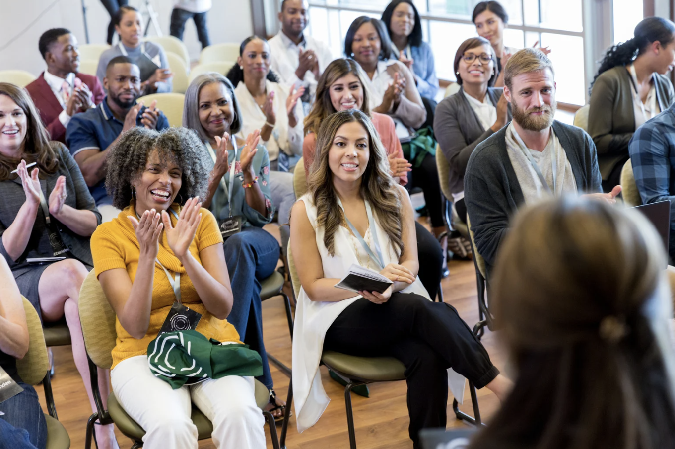 Group of diverse people sitting in an audience, applauding and smiling during an event in a well-lit conference room.