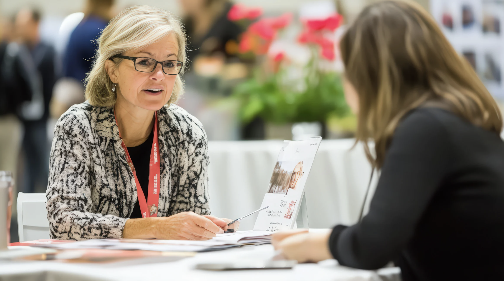 Two women having a conversation at a table in a busy indoor setting, one woman with blonde hair, glasses, and a patterned blazer, and the other with brown hair and a black blazer.