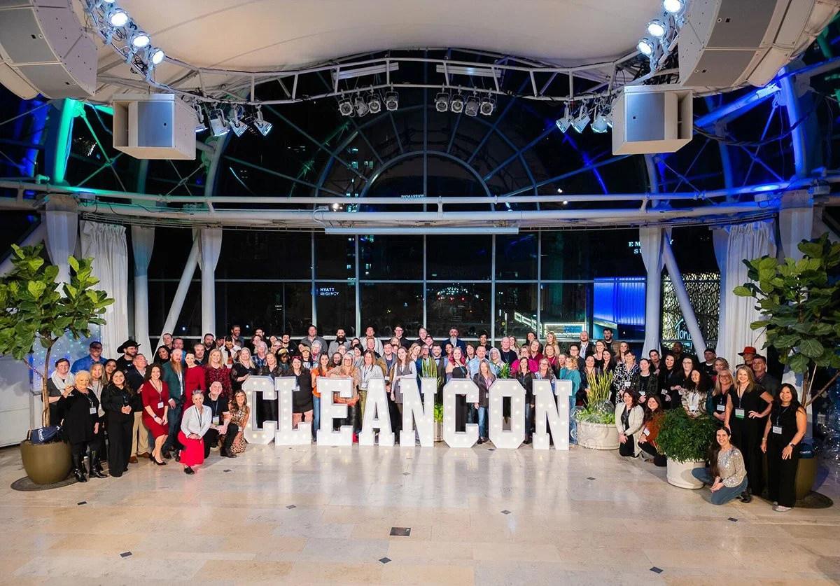 Group of people posing in front of large letter display spelling 'CLEANCON' at an indoor event venue with windows and plants.
