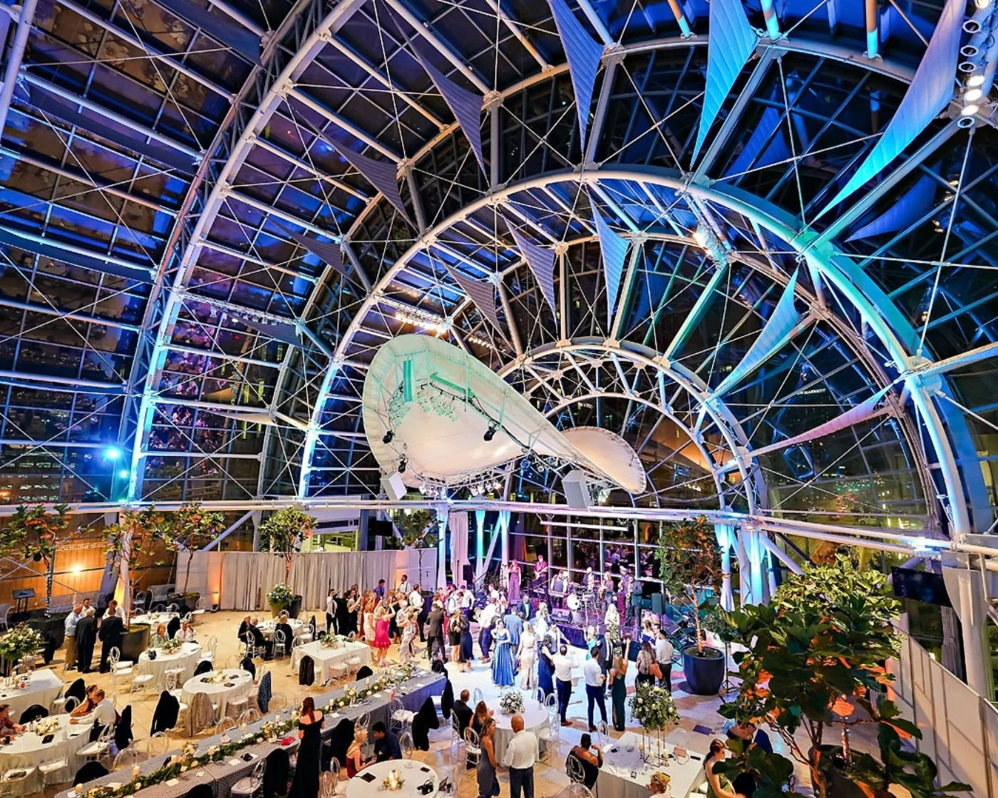 A lively indoor event at a glass-roofed venue with a modern, curved architecture, decorated with colorful lighting. Guests are mingling, dining at round tables, and a live band is performing on stage.