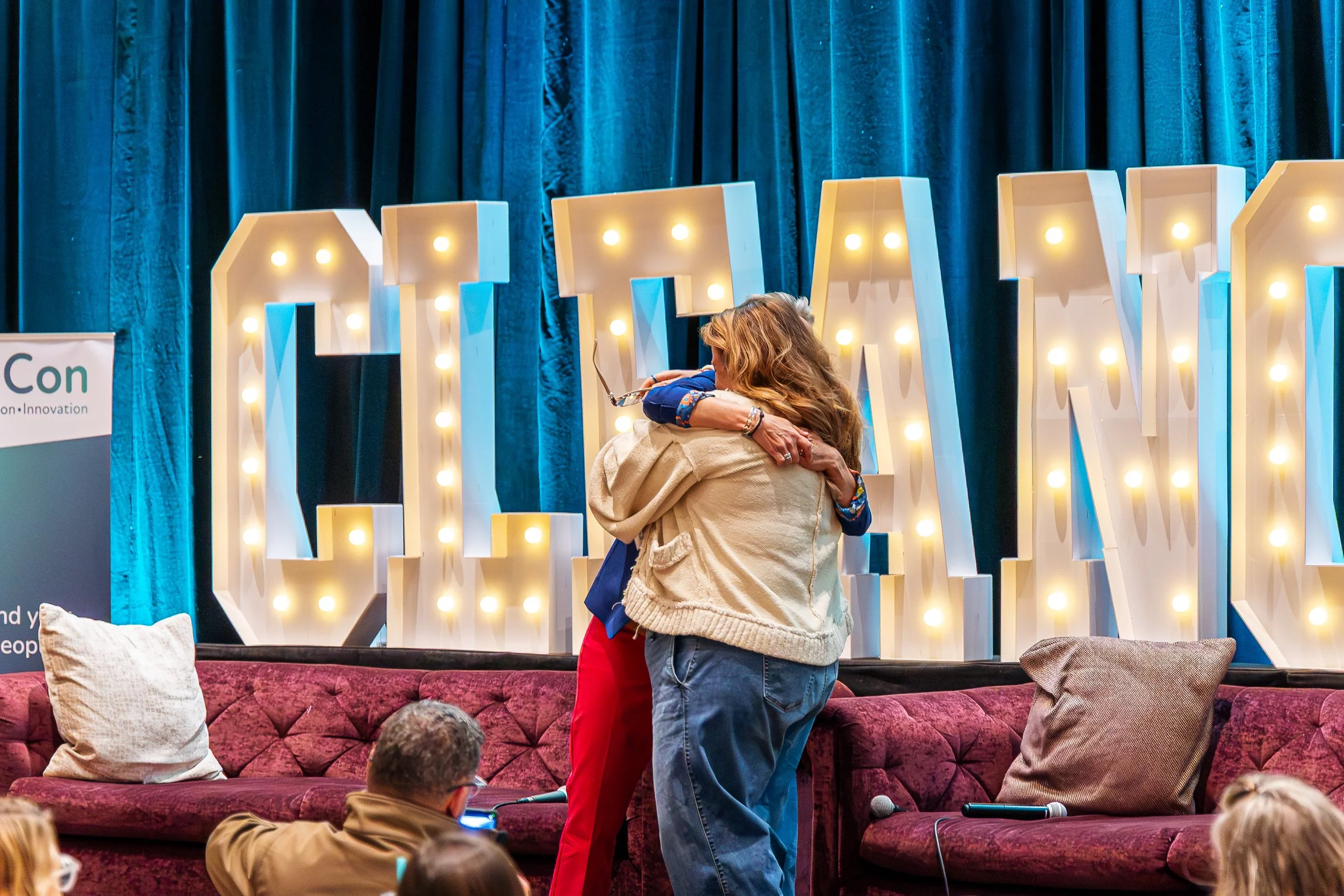 Two women hug on a stage with large illuminated letters behind them that spell out 'LAWS'. Stage curtains in the background and an audience in the foreground.