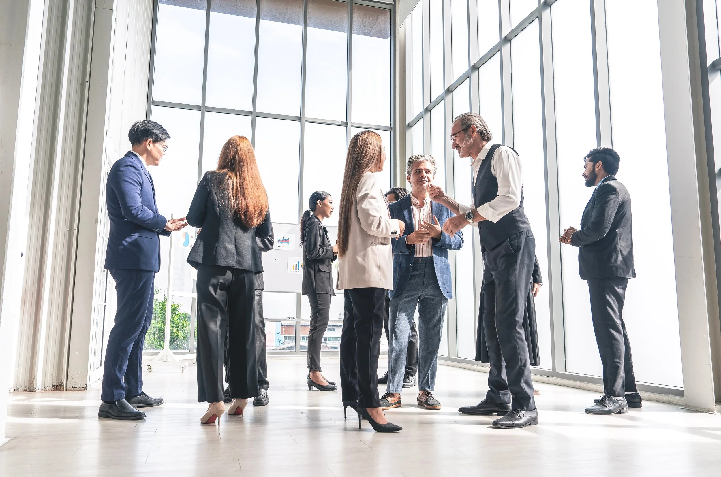 Business professionals in suits standing and talking in a modern, glass-walled office.
