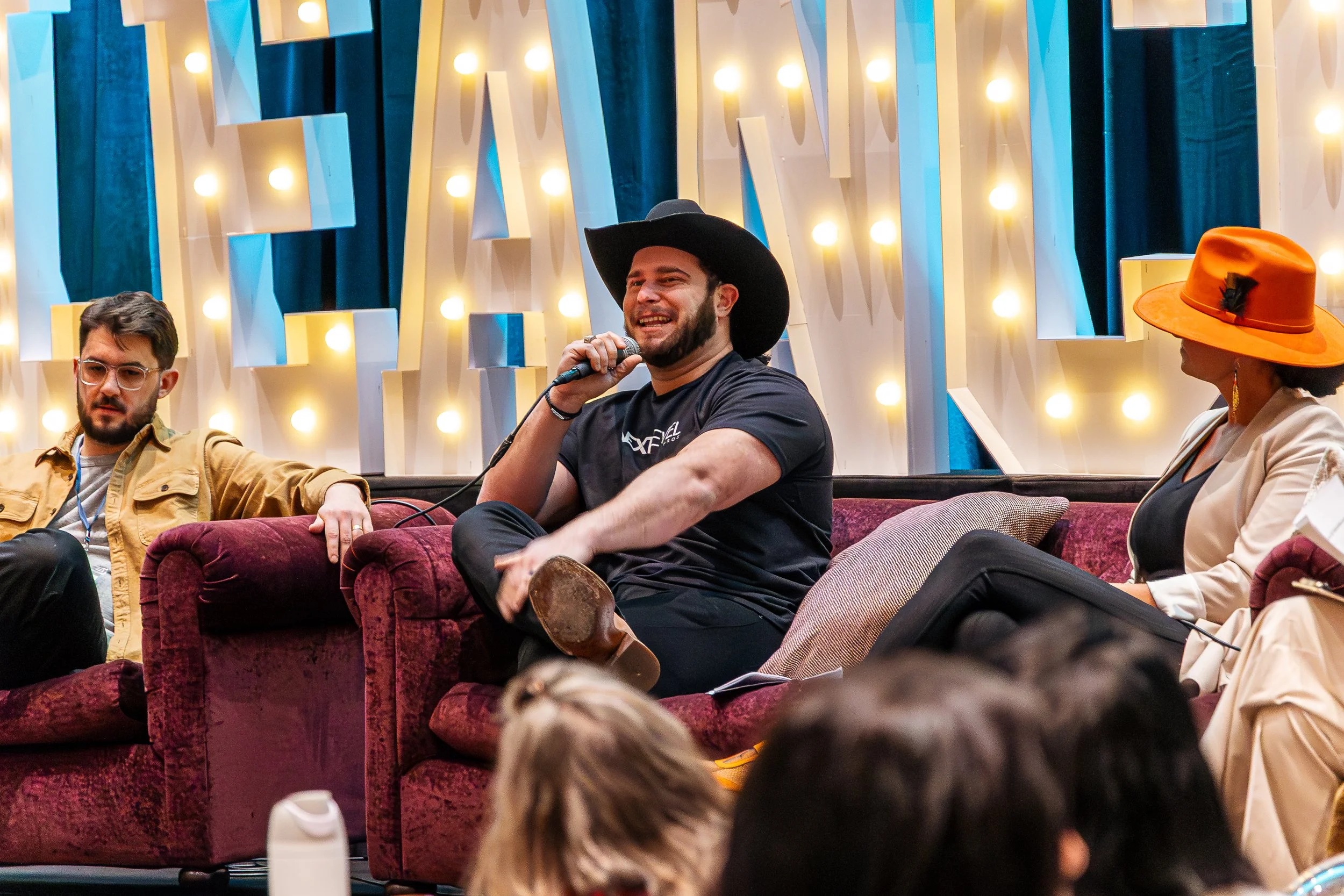 A man wearing a black cowboy hat smiling while speaking into a microphone during a panel discussion at an indoor event. Other panel members are sitting nearby, including a woman wearing a bright orange wide-brimmed hat and cream-colored blazer, and a man with glasses and a yellow jacket. The background features illuminated decorative wall panels.