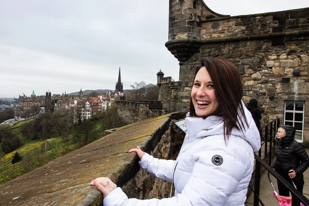 A woman with dark hair smiling and wearing a white puffer jacket, standing on a stone terrace of the Edinburgh castle with the Edinburgh, Scotland cityscape in the background.