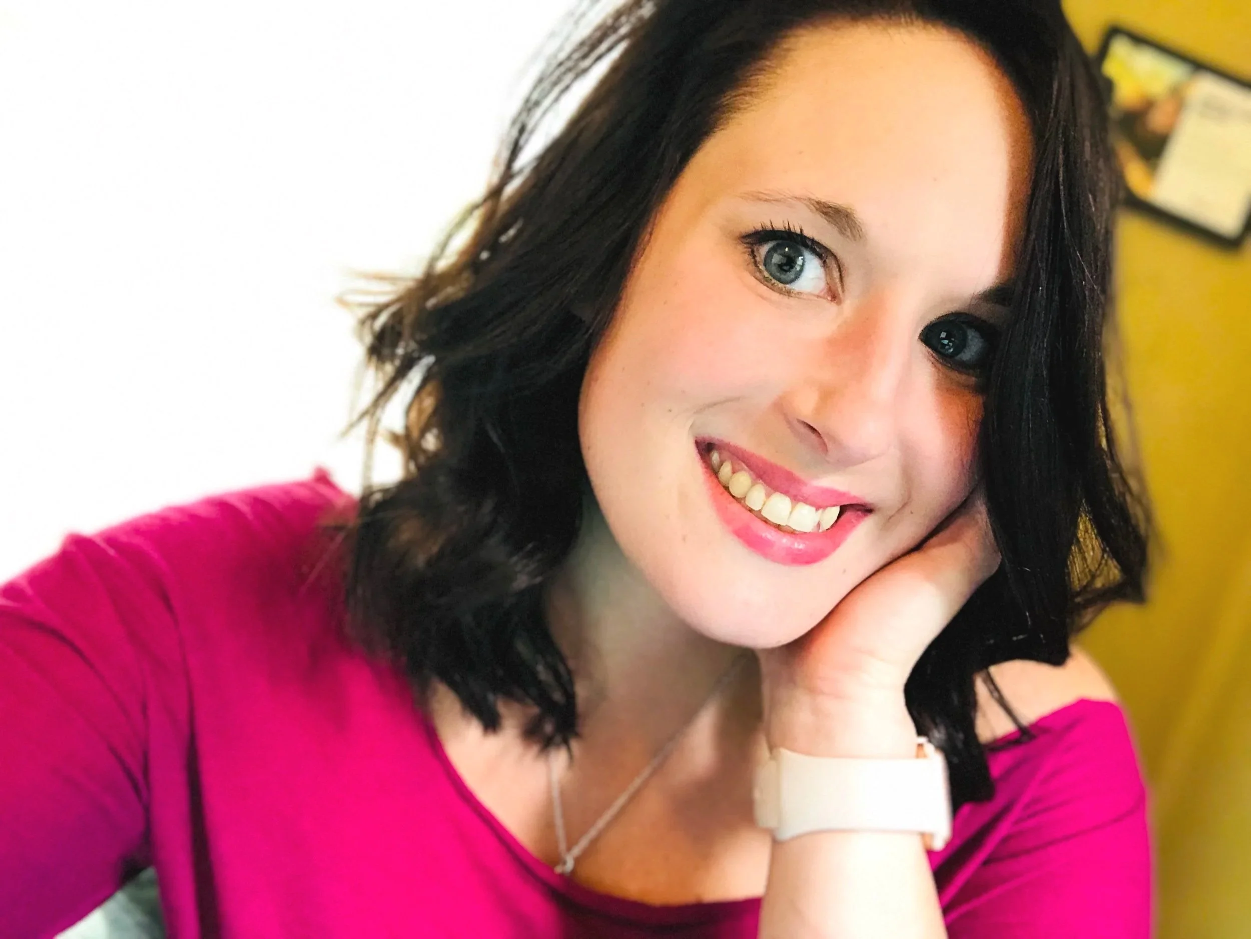 Close-up of a smiling woman with dark wavy hair, green eyes, pink lipstick, wearing a pink top, a silver necklace, and a light pink wristwatch, resting her chin on her hand.