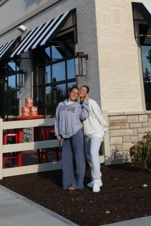 Two young women stand outside a building with a gray brick exterior, smiling and hugging each other. One wears a gray Notre Dame hoodie and loose pants, the other a white striped sweater and sneakers. Behind them, there is a beige fence with red chairs and two cans of Zima on a table. The building has large windows, black and white striped awnings, and outdoor lights.