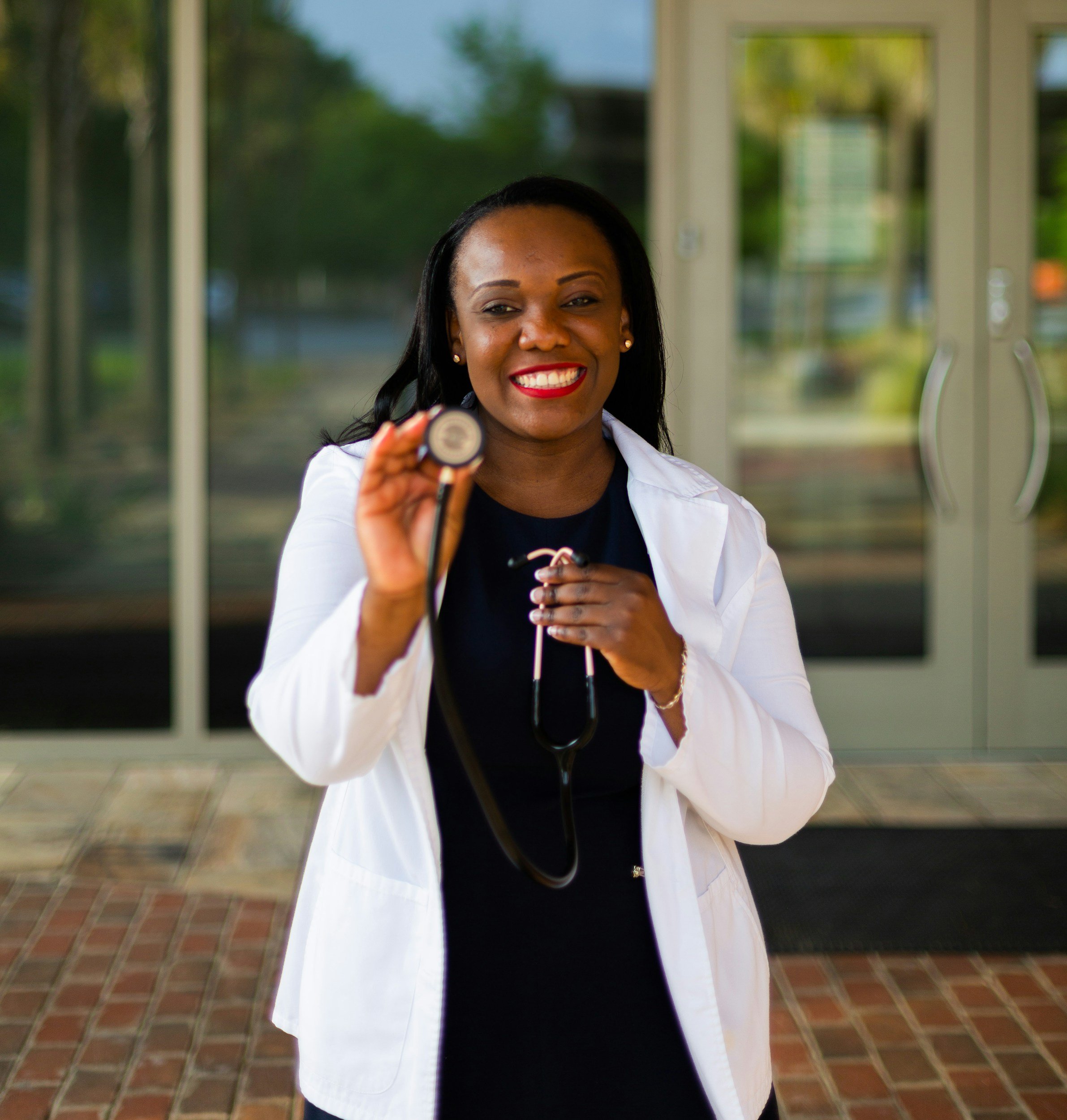 A smiling female healthcare professional in a white coat holding a stethoscope outside a building with glass doors.
