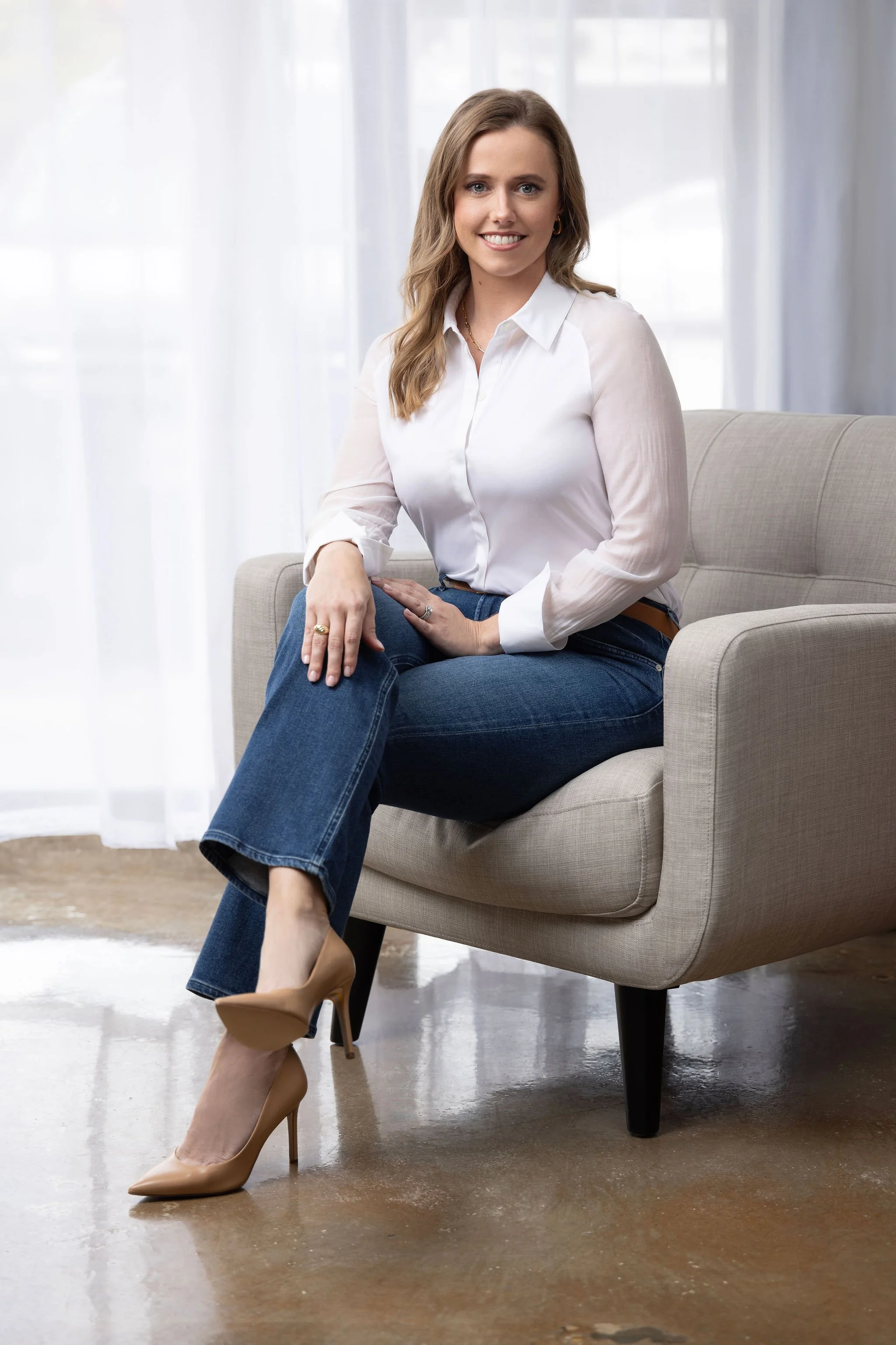 A woman with wavy light brown hair, wearing a white button-up shirt, blue jeans, and nude high heels, sitting on a beige armchair in a bright room with sheer white curtains.