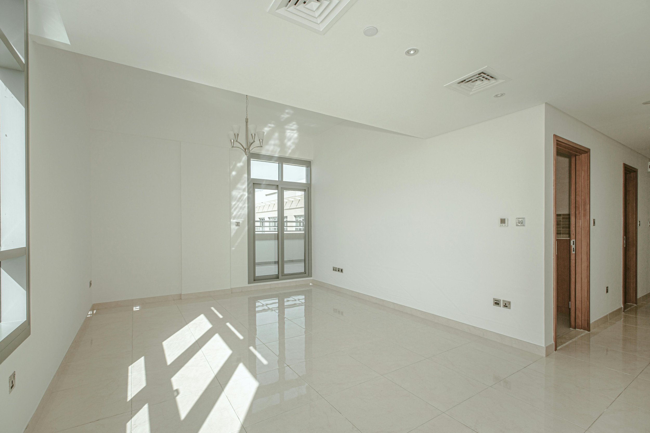 Empty room with white walls, large window with sliding door leading to a balcony, chandelier, and tiled floor.