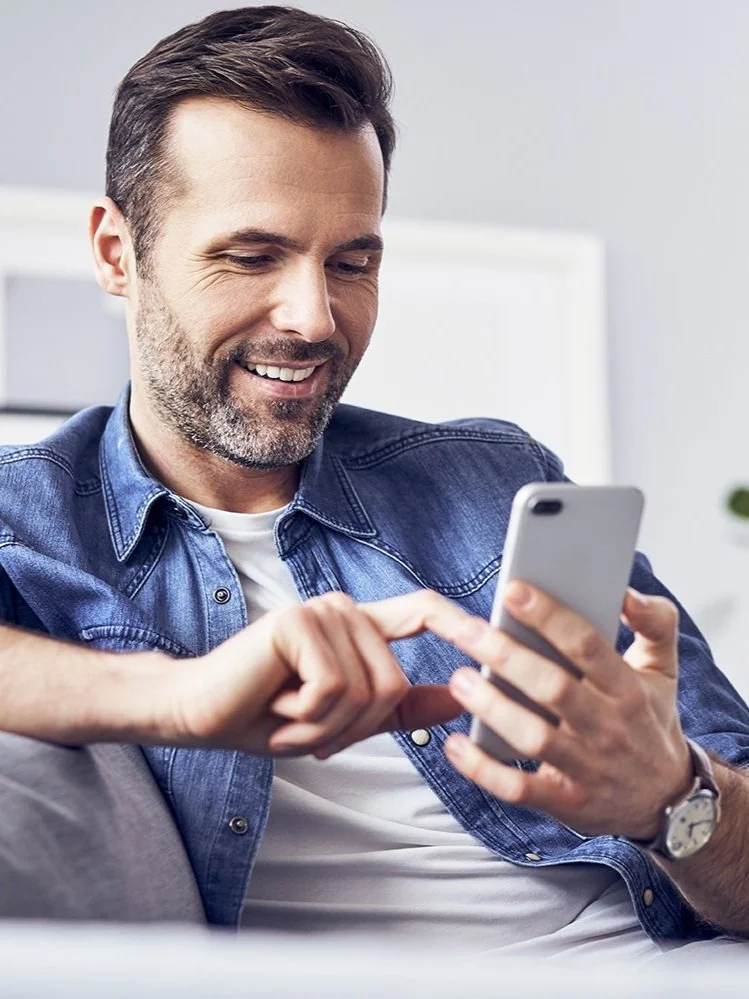 A man with a beard and short hair, wearing a denim shirt and a white t-shirt, is sitting on a couch looking at his smartphone and smiling.