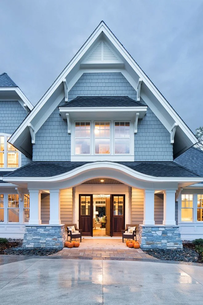 Front view of a modern house with blue and beige siding, a curved porch roof, and pumpkins outside the entrance during dusk.