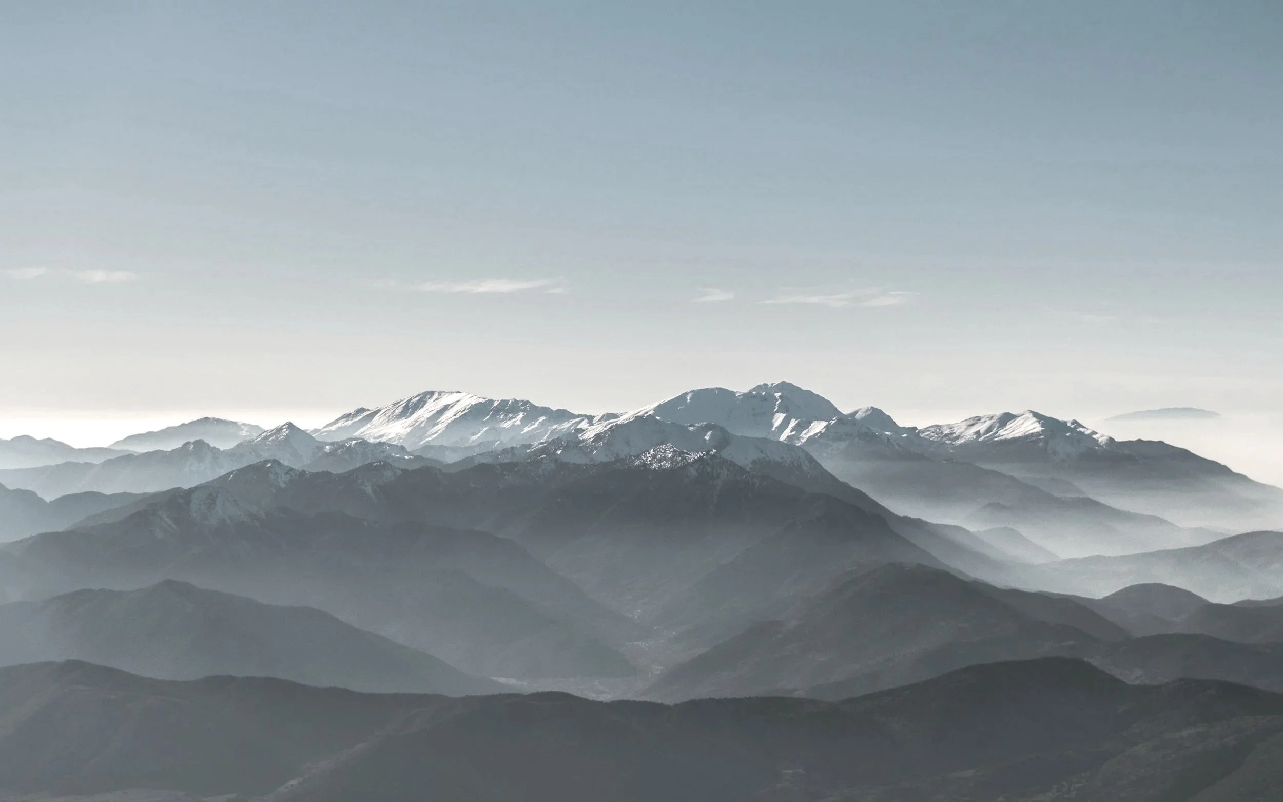 A landscape of multiple mountain ranges with snow-capped peaks, layered in shades of gray and blue, under a pale sky.