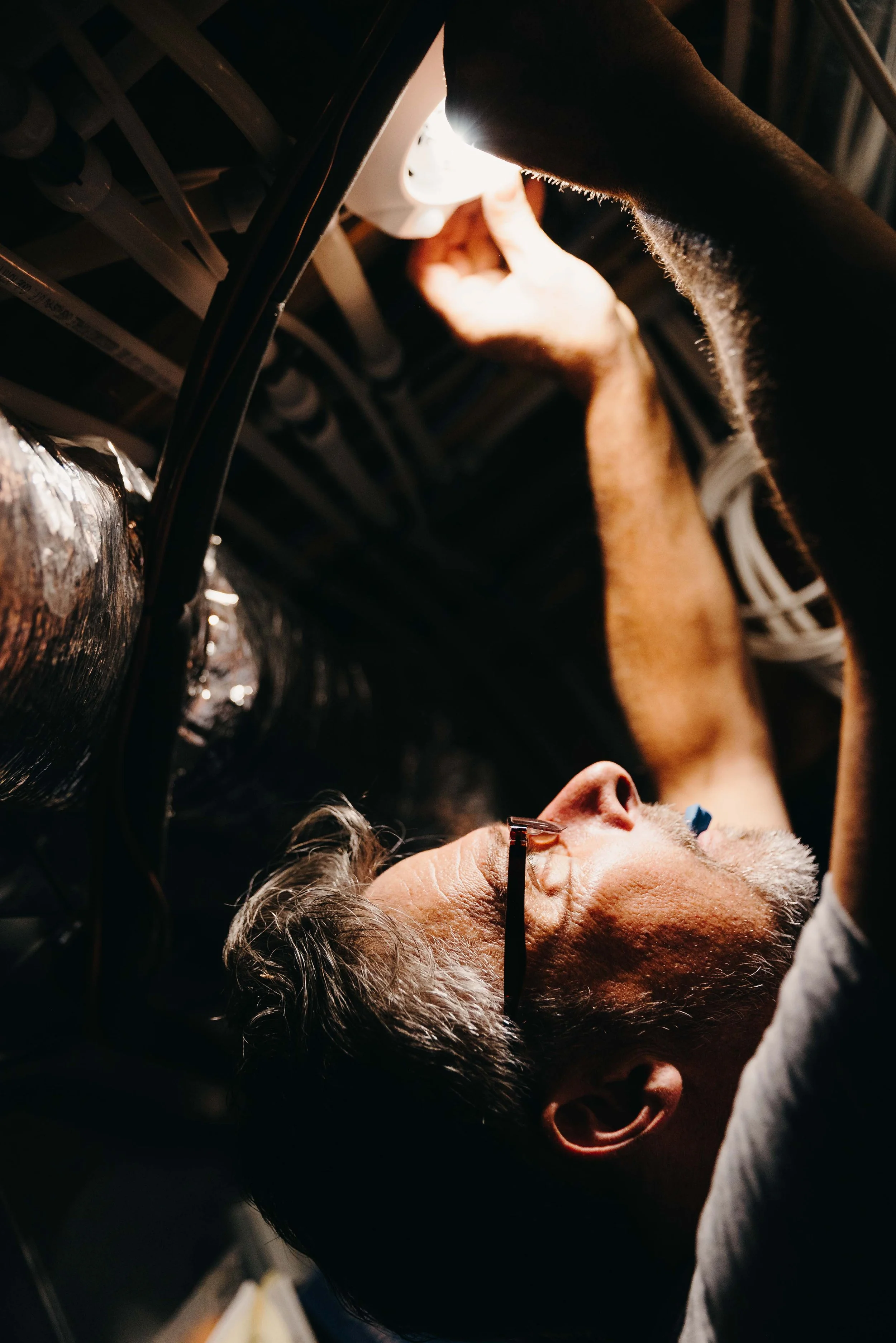 Andy Ferguson working on electrical wiring or equipment, lying on his back under a structure with exposed pipes and wires, illuminated by a bright light.