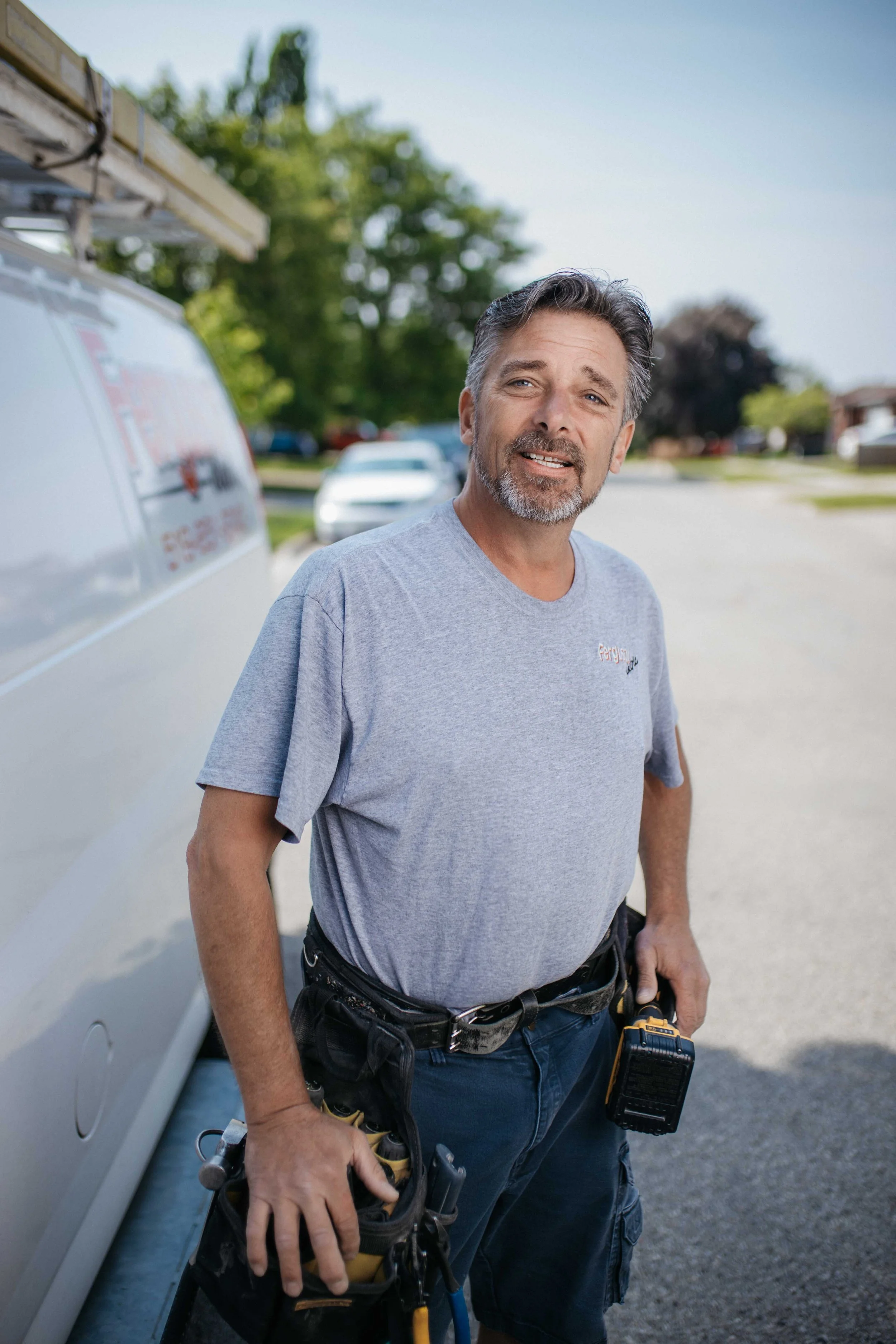 Andy Ferguson wearing a gray T-shirt and work tool belt, standing outdoors next to a utility van, holding tools, with a suburban street in the background.