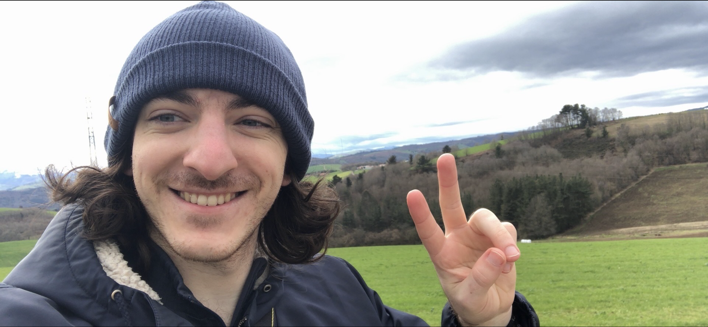 A smiling man with long curly hair, wearing a gray beanie and a dark jacket, taking a selfie outdoors in a rural landscape with green rolling hills and cloudy sky in the background.