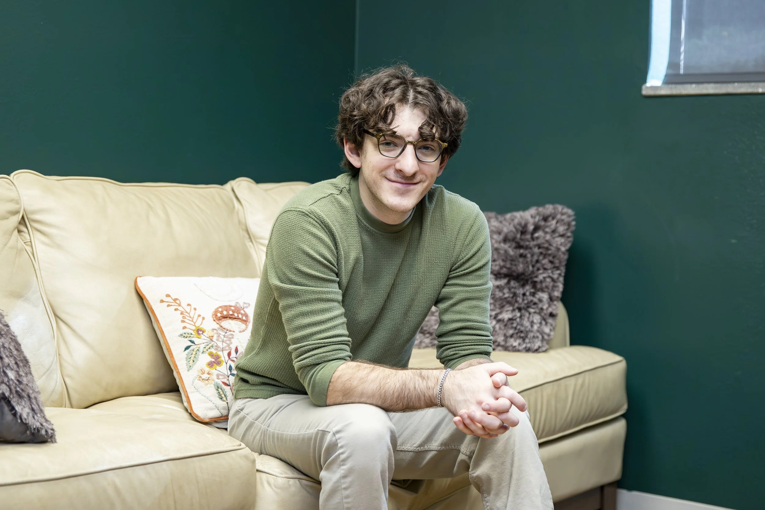 A young man with curly hair and glasses sitting on a beige couch, wearing a green long sleeve shirt and khaki pants, smiling at the camera in a room with dark green walls.