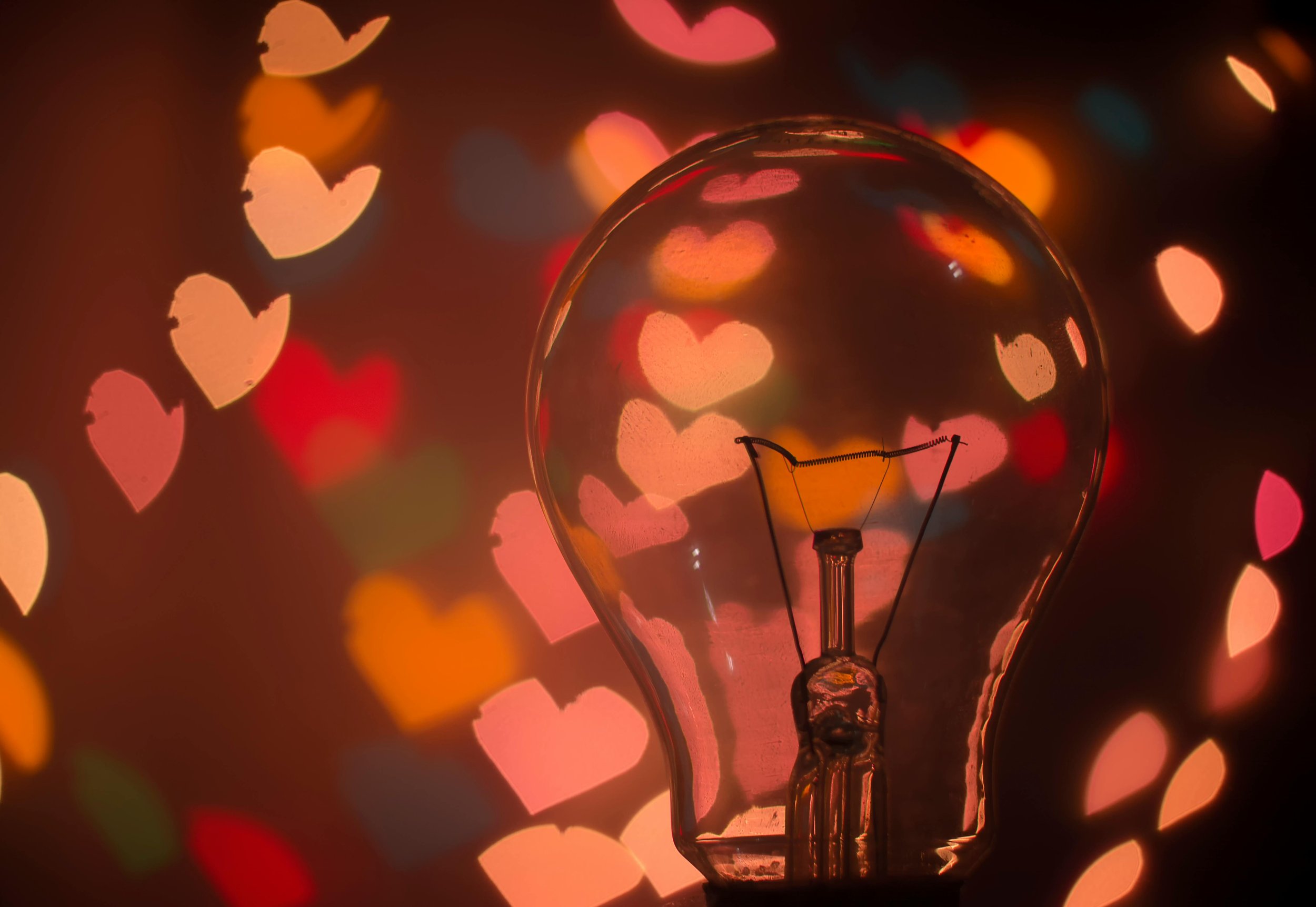 A close-up of an incandescent light bulb with a background of colorful heart-shaped bokeh lights in red, pink, orange, yellow, and teal.