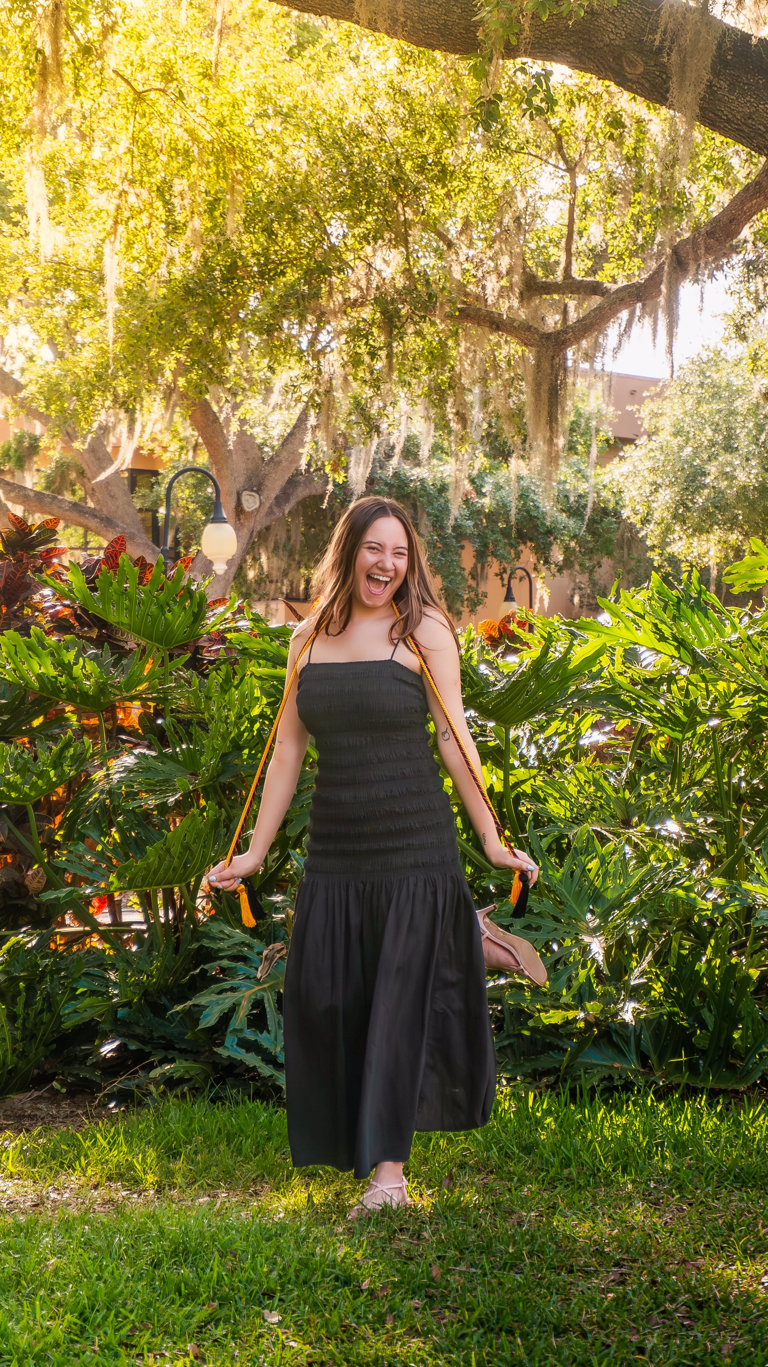 A woman in a black dress standing outdoors with lush green plants and trees, smiling and laughing.