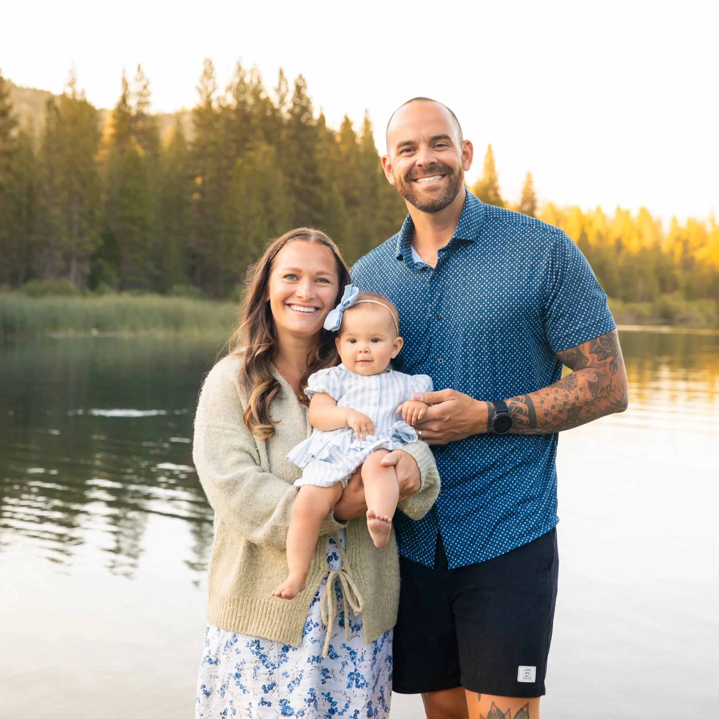 A family of three standing by a lake with trees in the background, during sunset. The woman is holding a baby girl, and the man is smiling next to them.