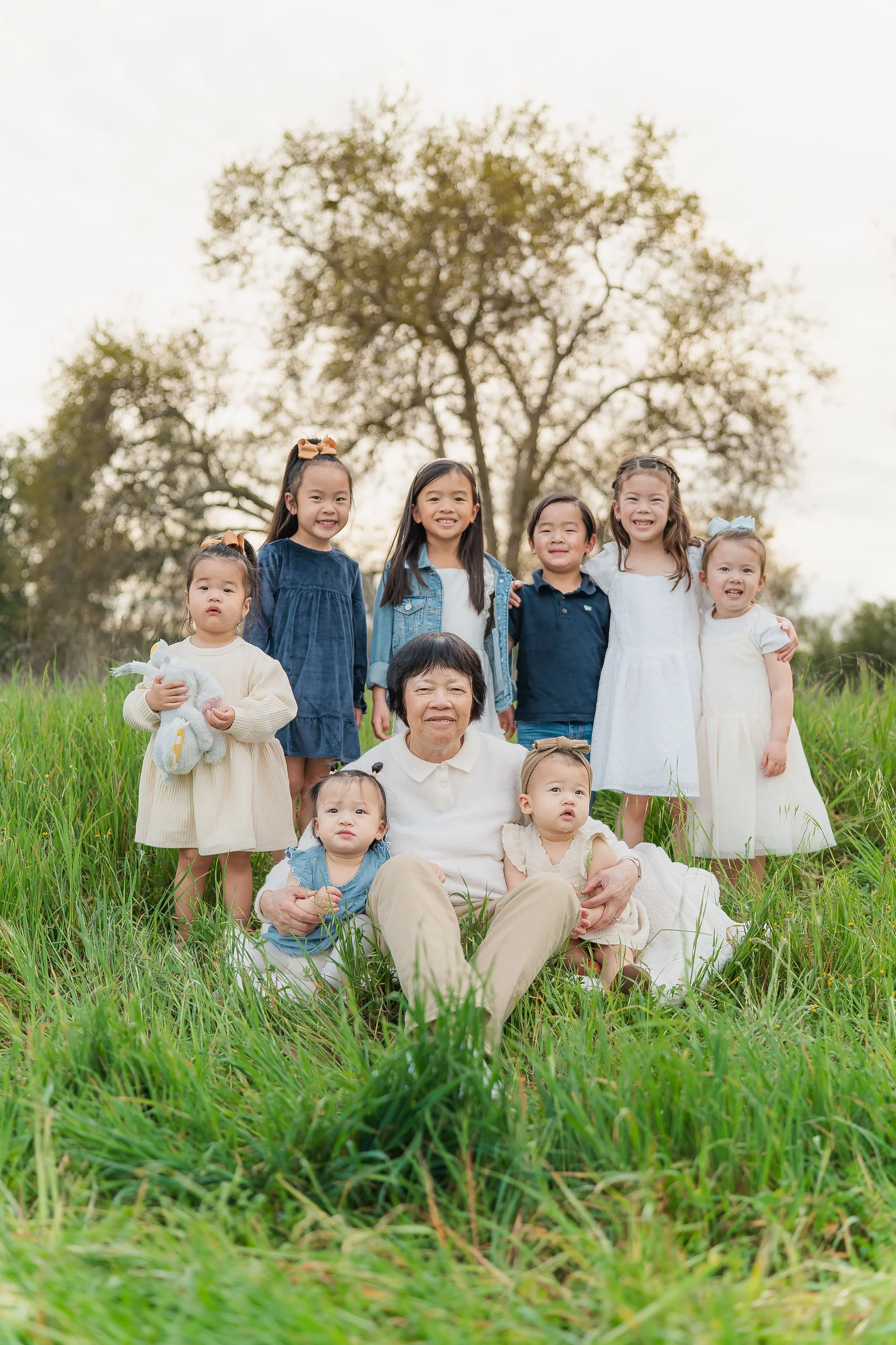 A group of nine children and an elderly woman sitting on grass outdoors, with trees and a cloudy sky in the background.