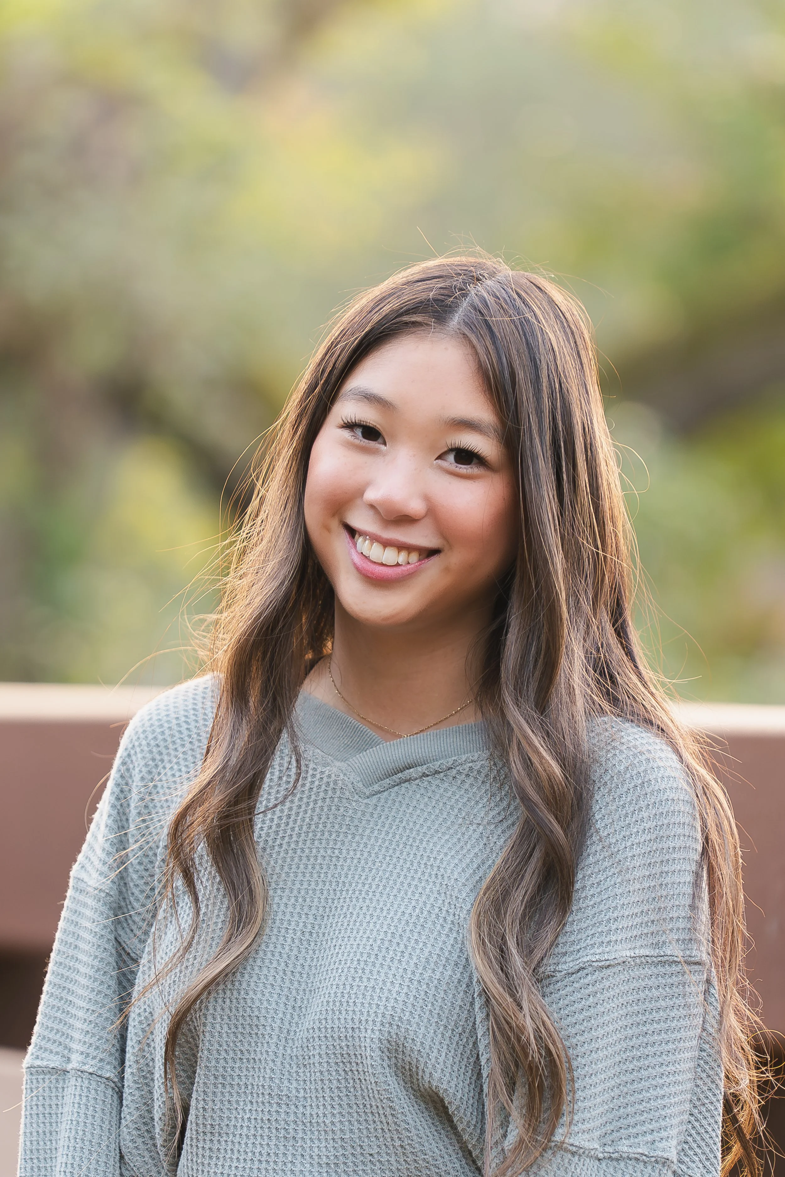 A young woman with long wavy hair smiling outdoors with a blurred natural background.