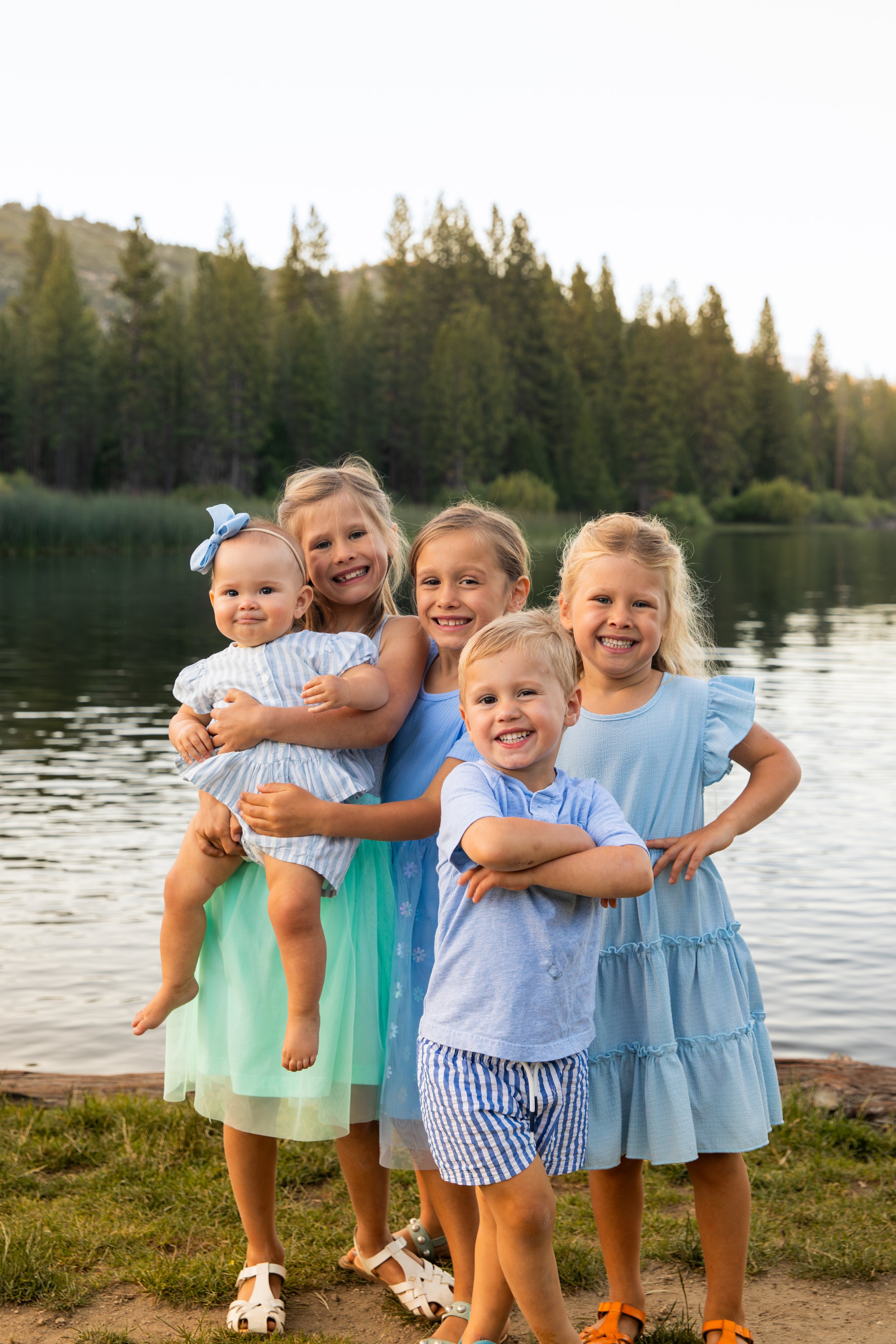 Six children, four girls and two boys, smiling and posing together outdoors near a lake with a forested background.