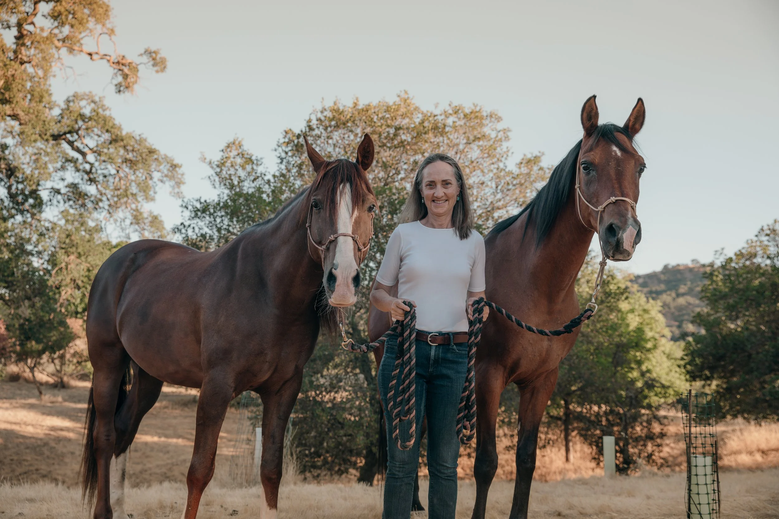 A woman standing outdoors holding the reins of two brown horses, with trees and a hillside in the background.