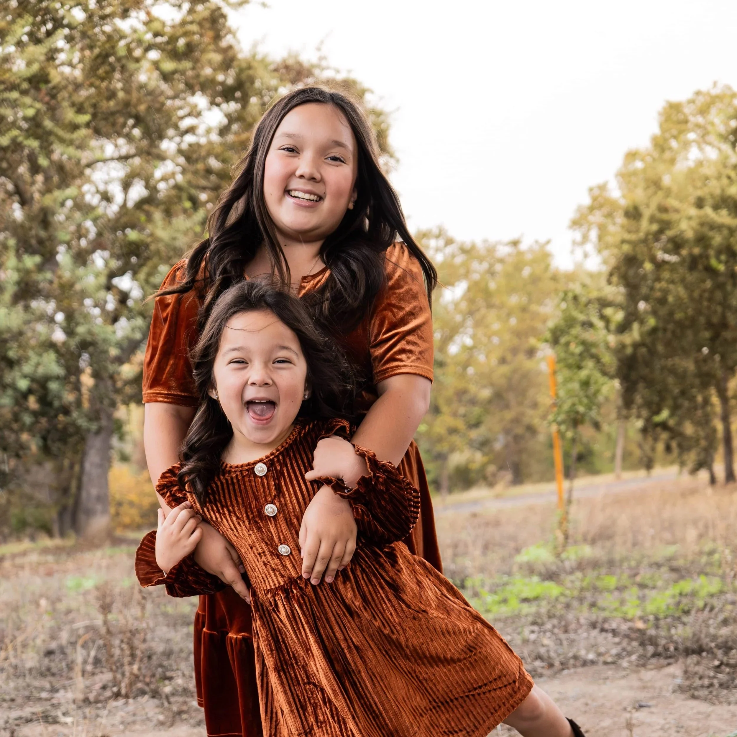Two young girls, one older and one younger, smiling and playing outdoors in an autumn setting with trees and open field in the background.