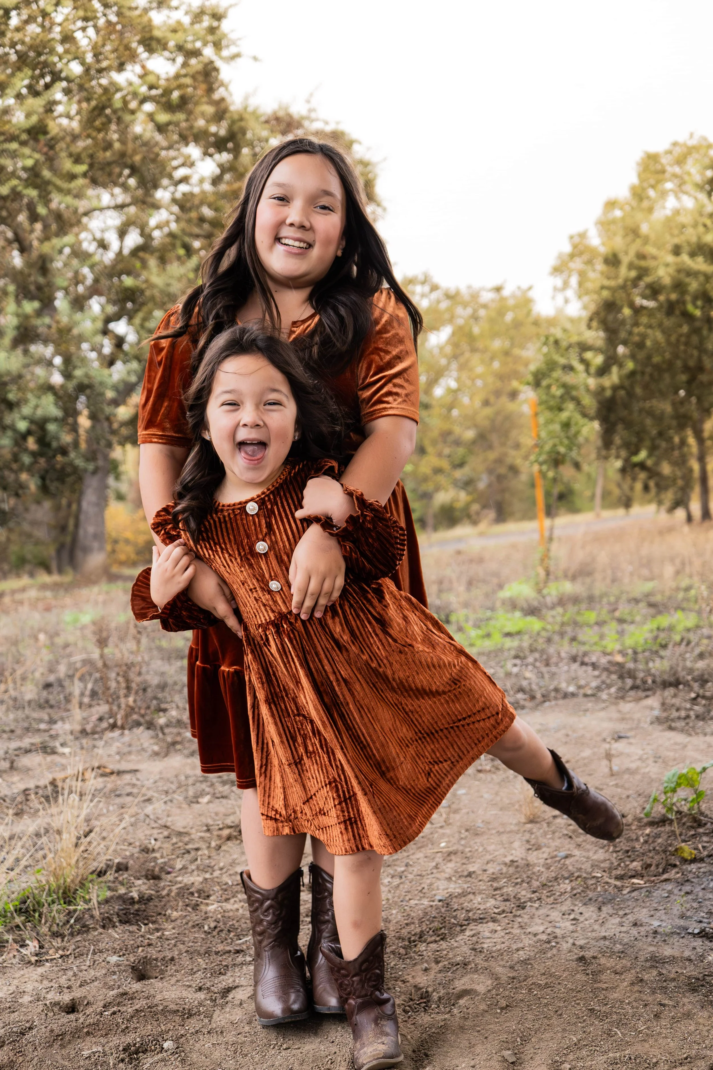 Two young girls with long dark hair, wearing matching rust-colored velvet dresses and cowboy boots, standing outdoors in a field with trees in the background, smiling and playing.