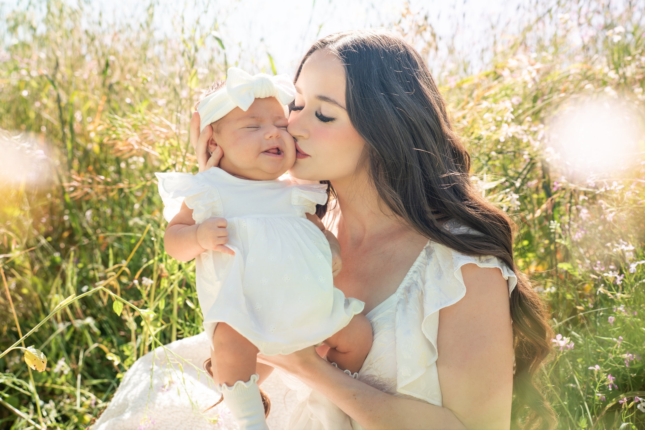 A woman holding a young girl in a grassy outdoor setting with sunlight, both wearing white dresses. The woman is kissing the girl's cheek while the girl has her eyes closed and a slight smile.