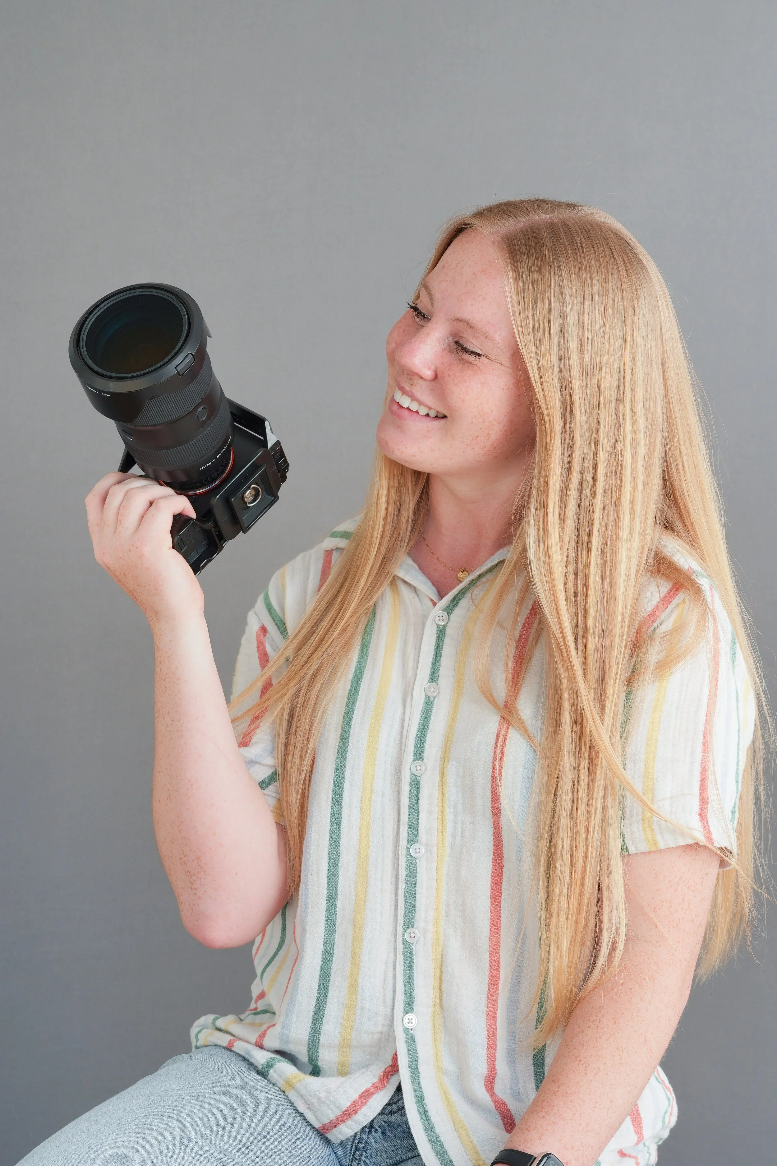 A young woman with long blonde hair, wearing a striped shirt, smiling and holding a camera in her hand against a plain gray background.