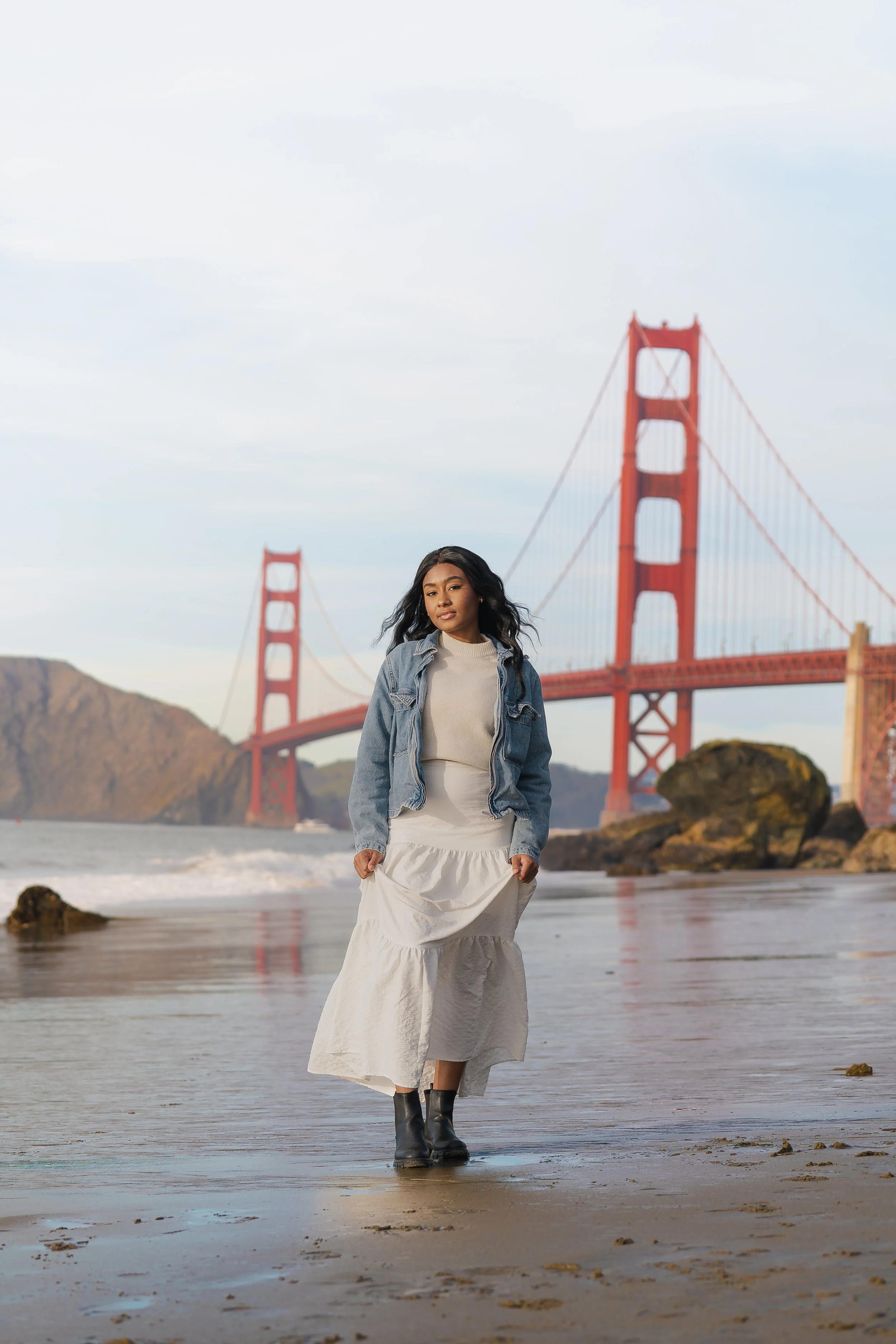 A woman walking on a beach near the Golden Gate Bridge in San Francisco, wearing a denim jacket, white dress, and black boots.