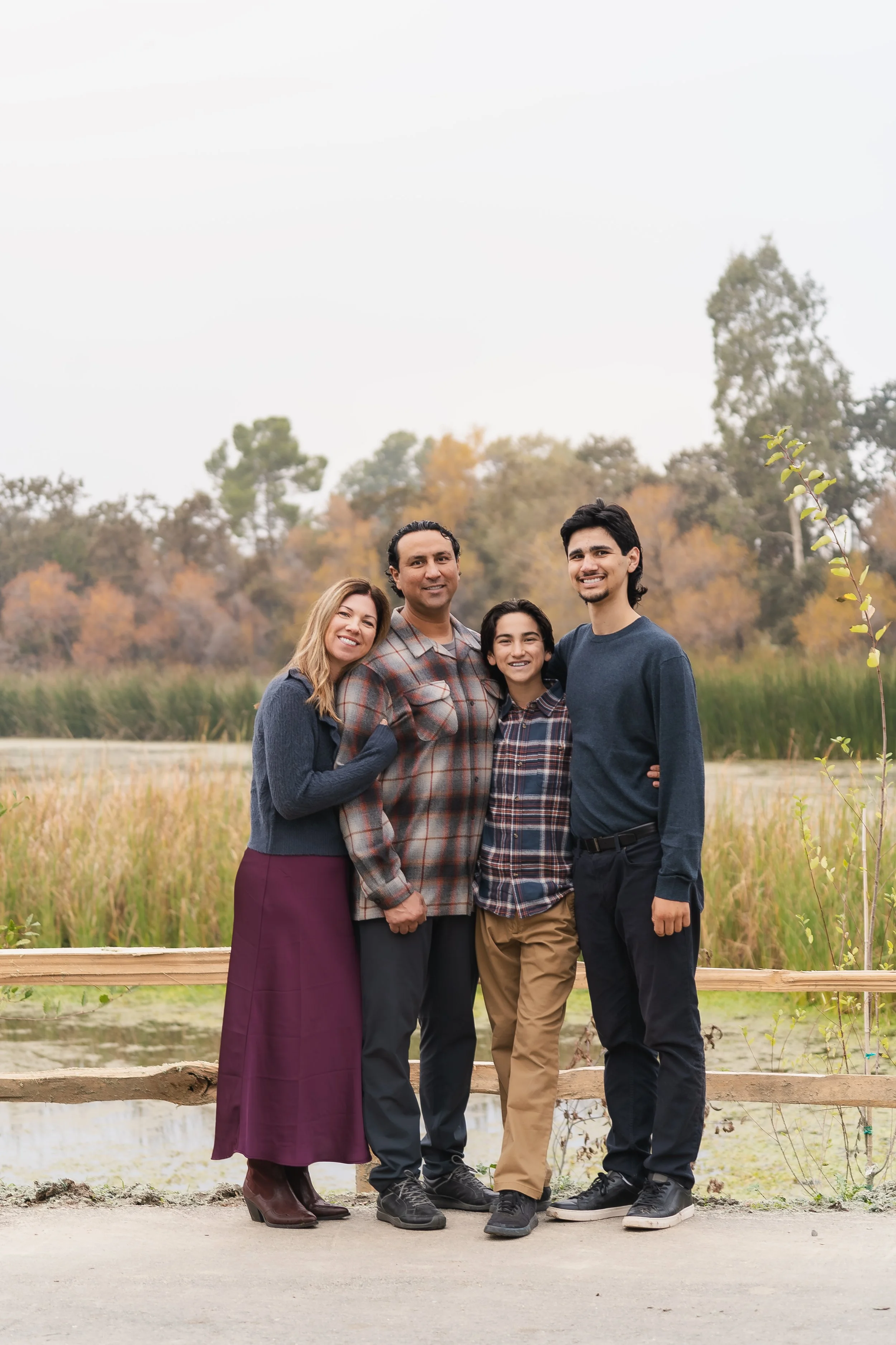 A family of four standing outdoors near a wooden fence, smiling, with trees and tall grass in the background.