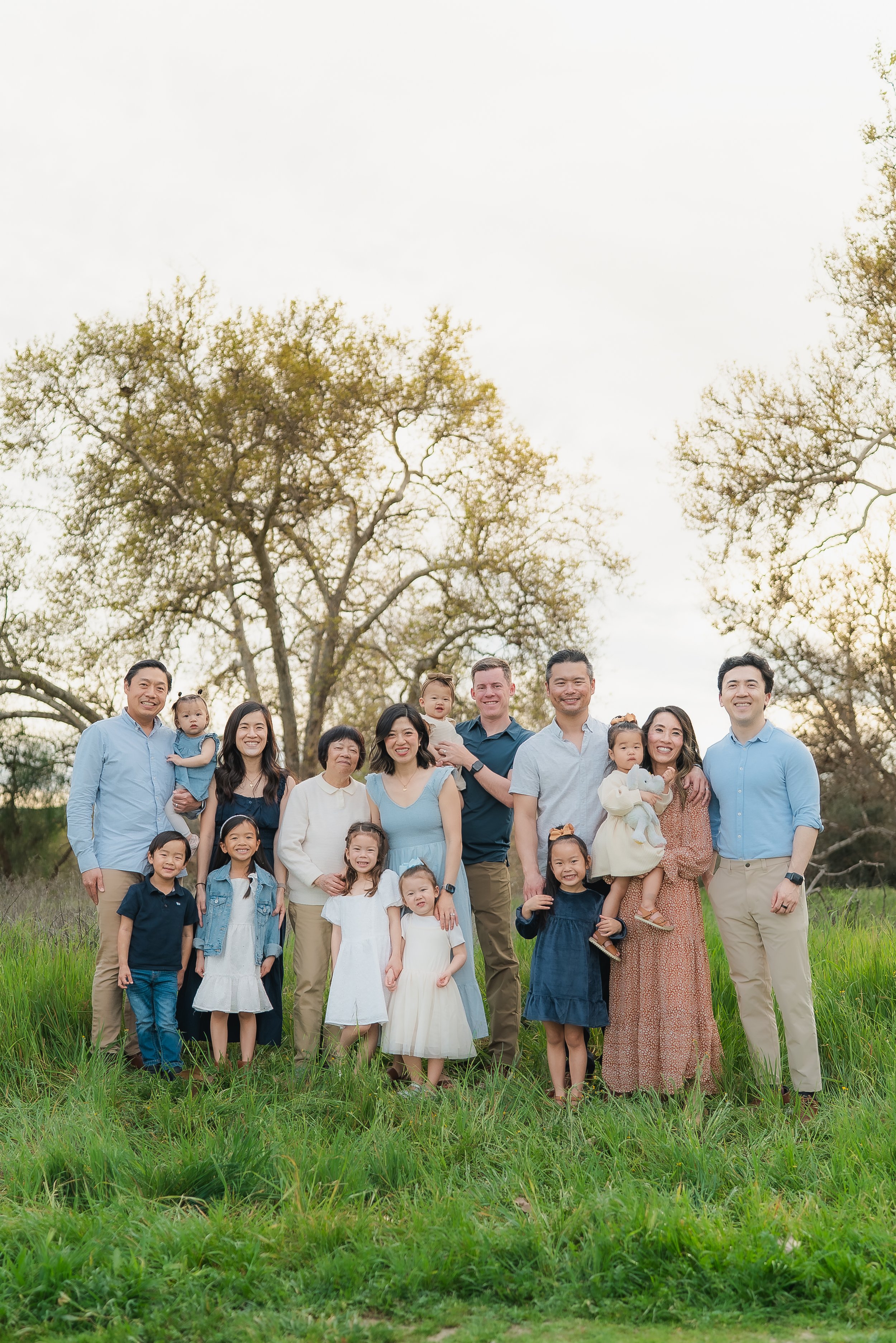 Family group photo outdoors with adults and children, standing on grass, trees in the background, during daytime.