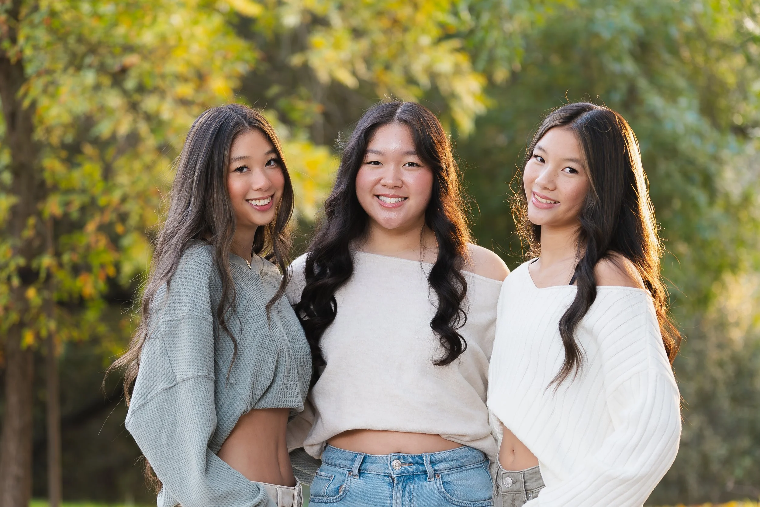 Three young women standing outdoors in a park with trees and greenery in the background, smiling at the camera.