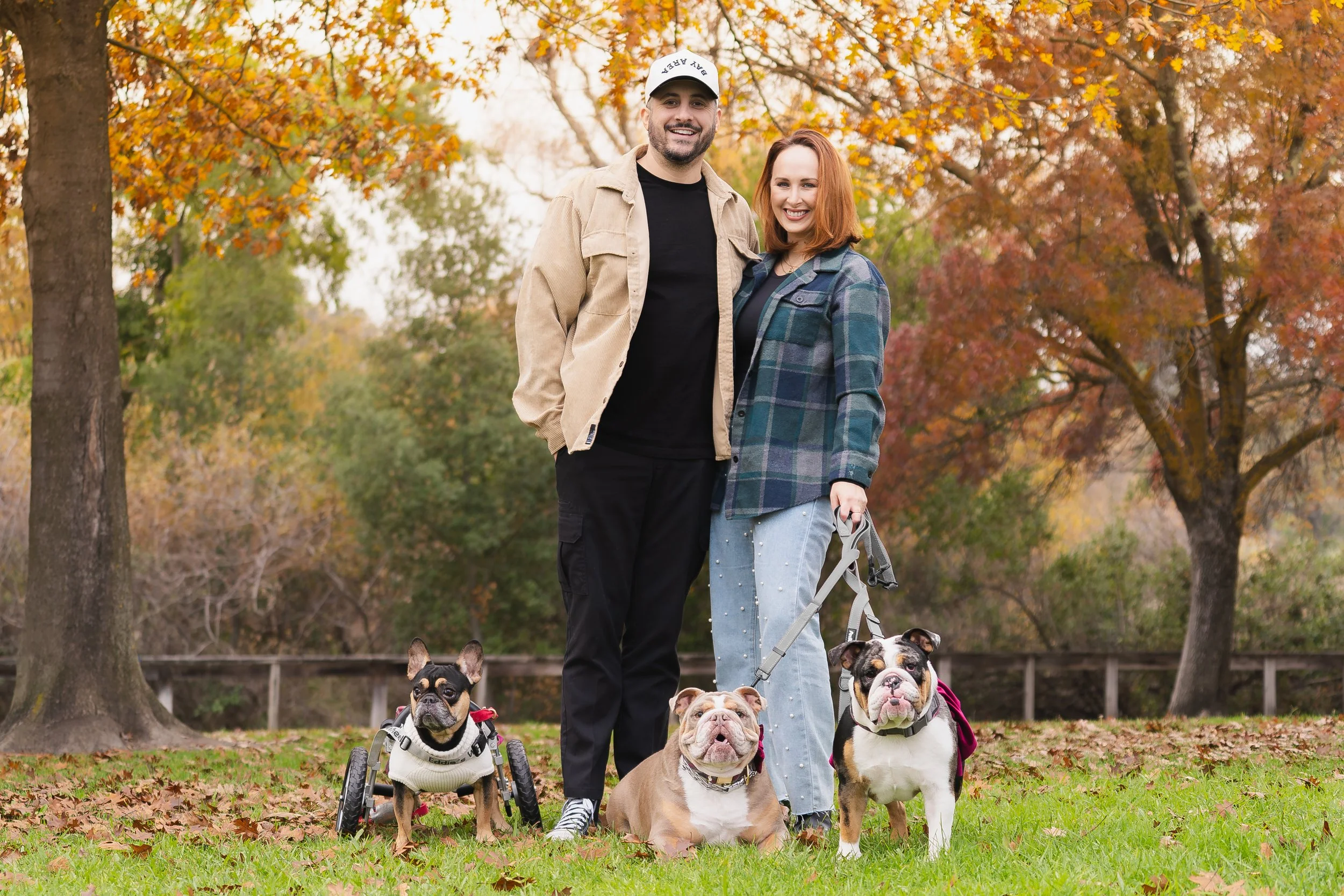 A smiling couple standing outdoors in a park during fall, surrounded by trees with colorful autumn leaves, with three dogs including one using a wheelchair.