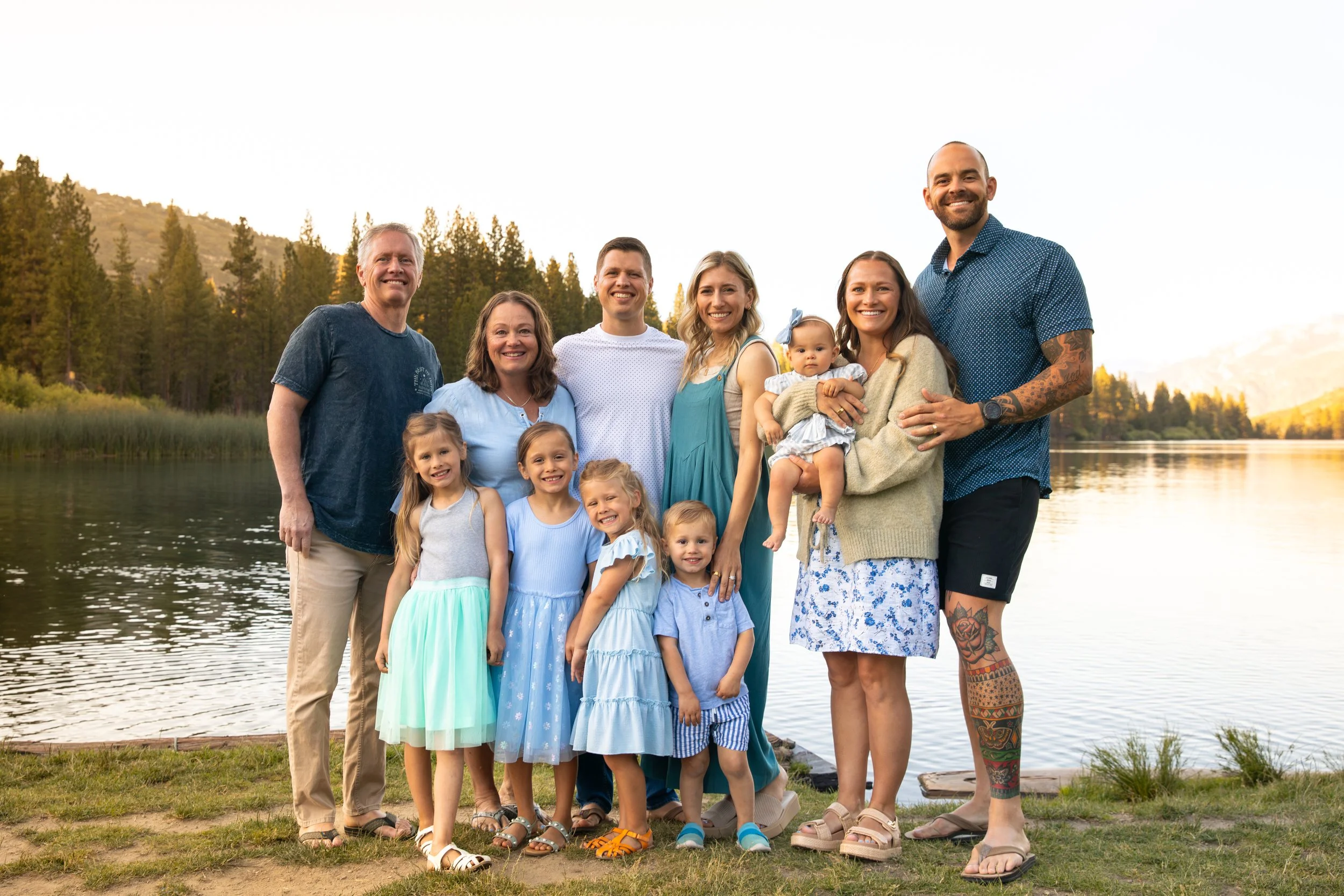 A large family group with seven adults and five children, standing outdoors by a lake with trees and mountains in the background, during sunset.