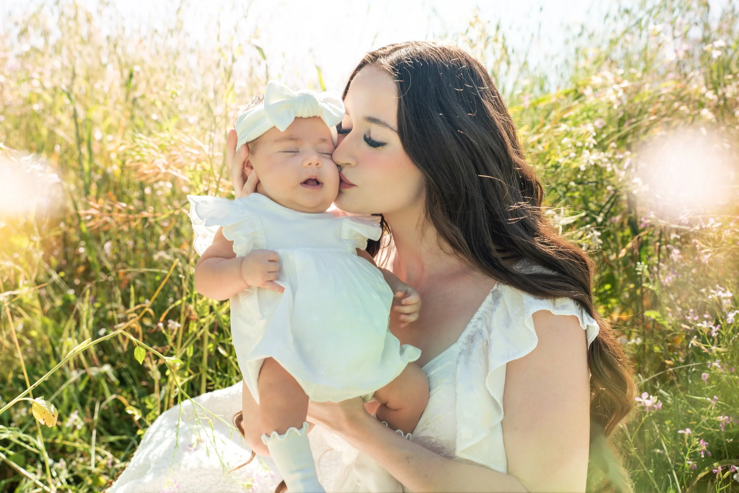 A woman holding a baby girl outdoors in a grassy field, with the woman kissing the baby's cheek, both dressed in white, during daytime.