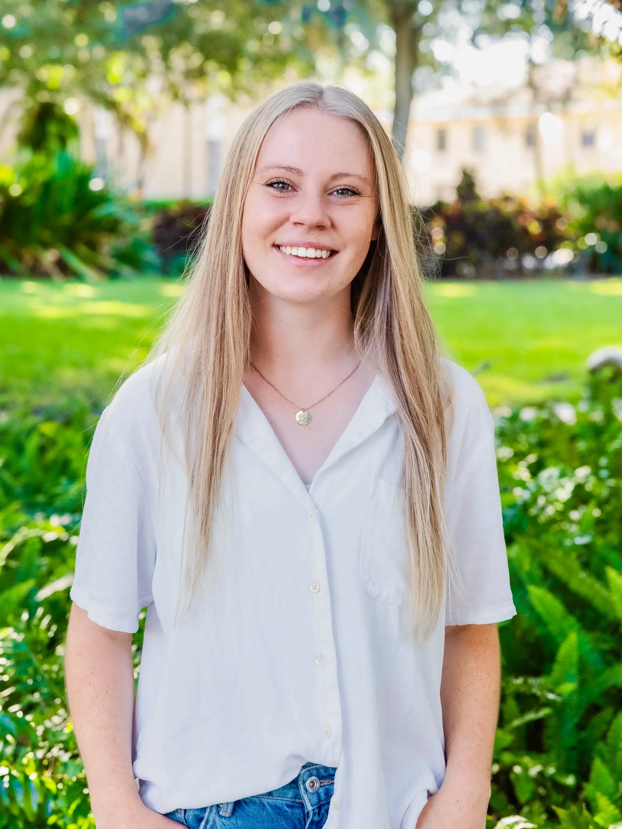 Smiling young woman with long blonde hair wearing a white button-up shirt and a necklace, standing outdoors in a lush green park.