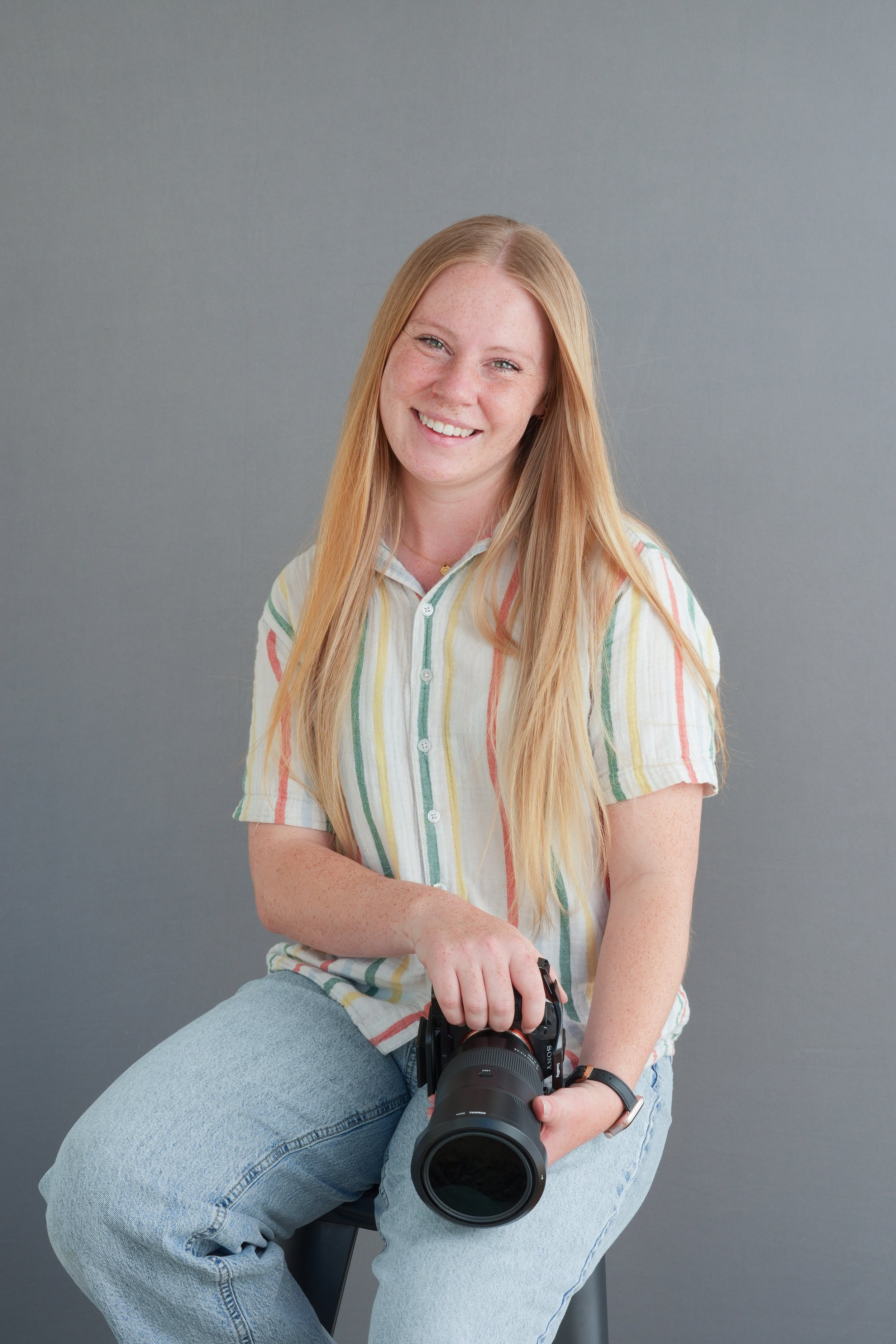 Young woman with long red hair, wearing a striped shirt, holding a camera, sitting on a stool against a gray background.
