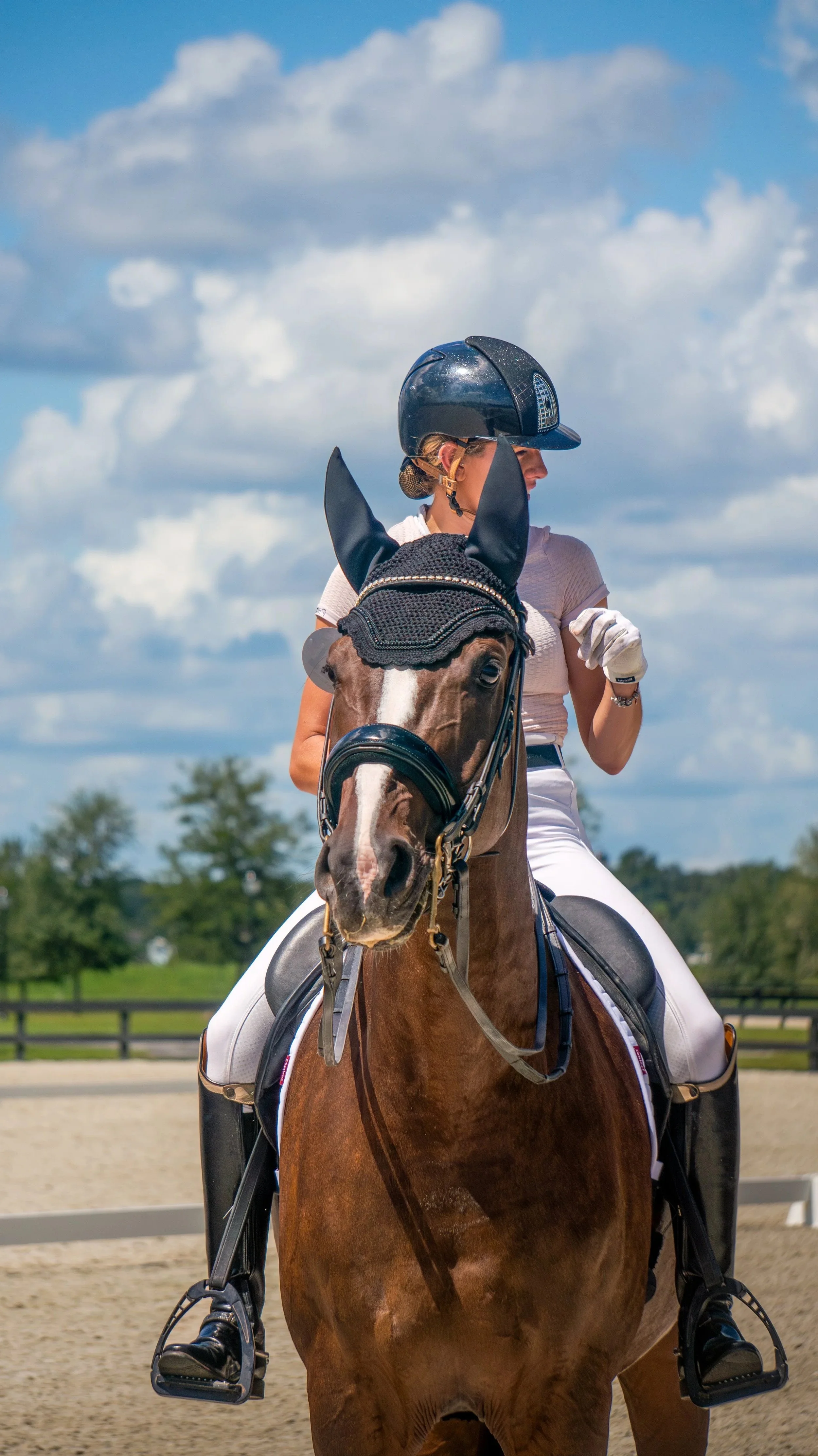 A woman riding a brown horse on an outdoor riding arena with a blue sky and scattered clouds in the background.