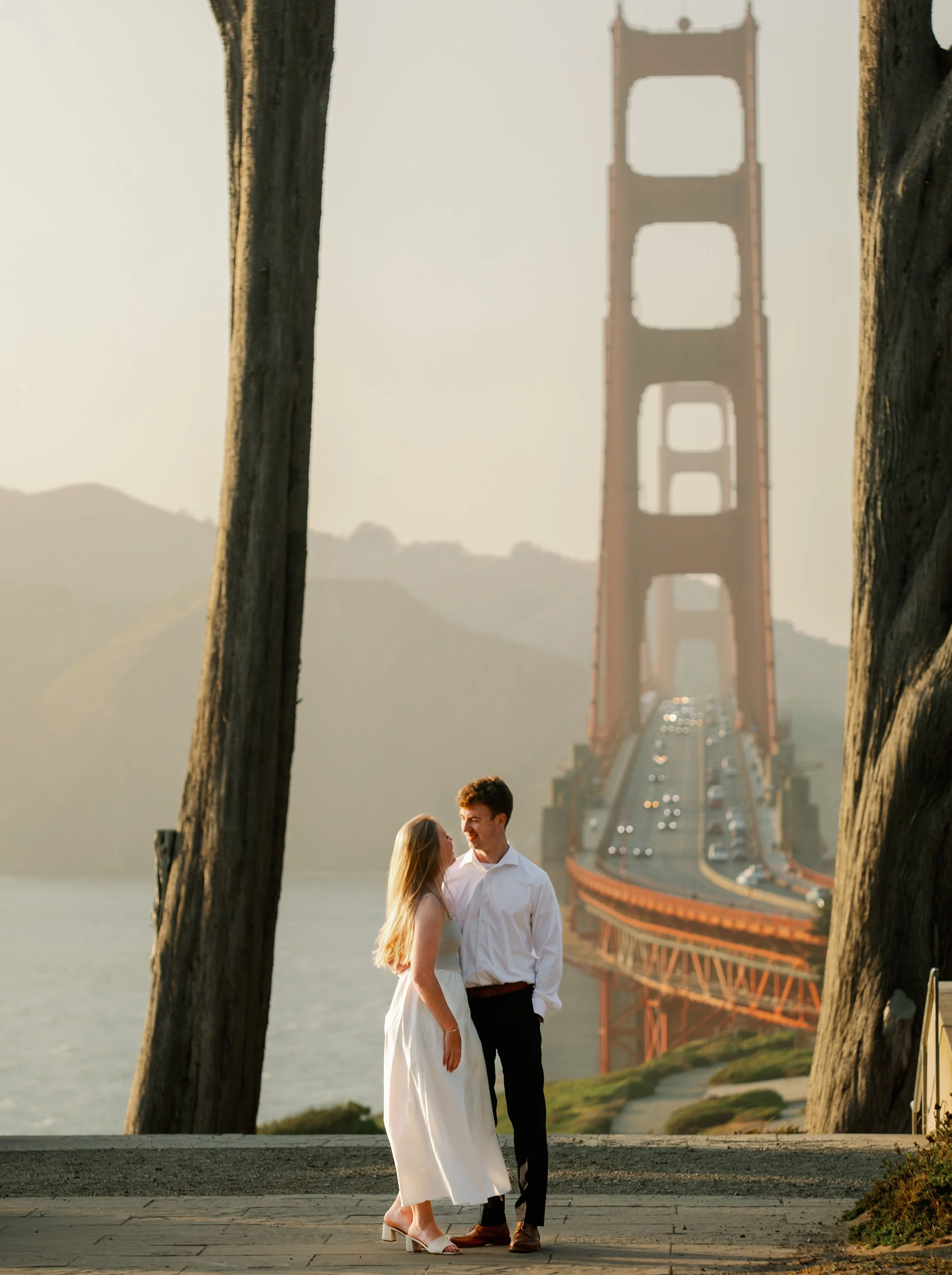 A couple in wedding attire standing together near the Golden Gate Bridge, with the bridge and its traffic in the background during sunset.