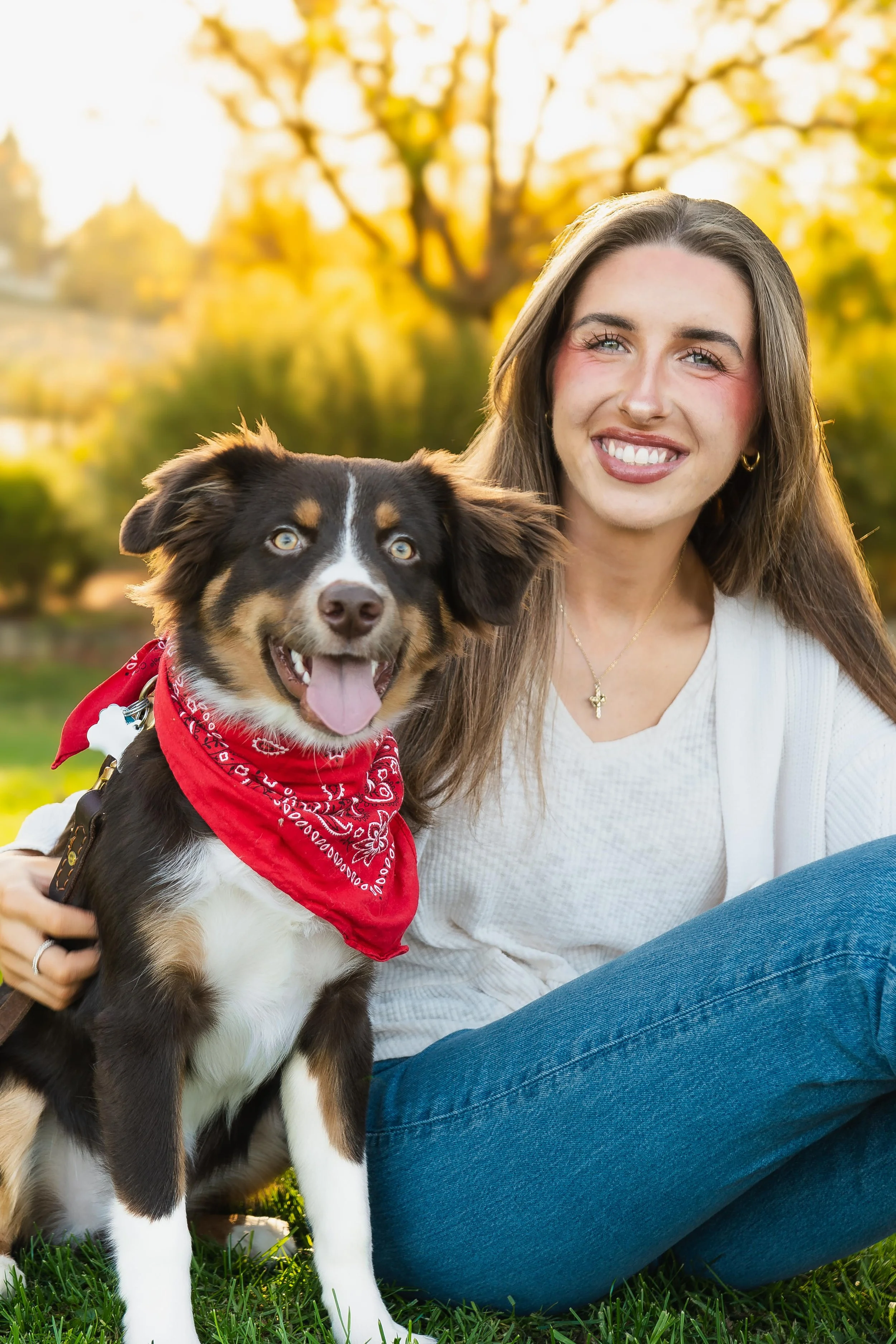 A young woman and her dog sitting on the grass in a park during golden hour with autumn trees in the background.