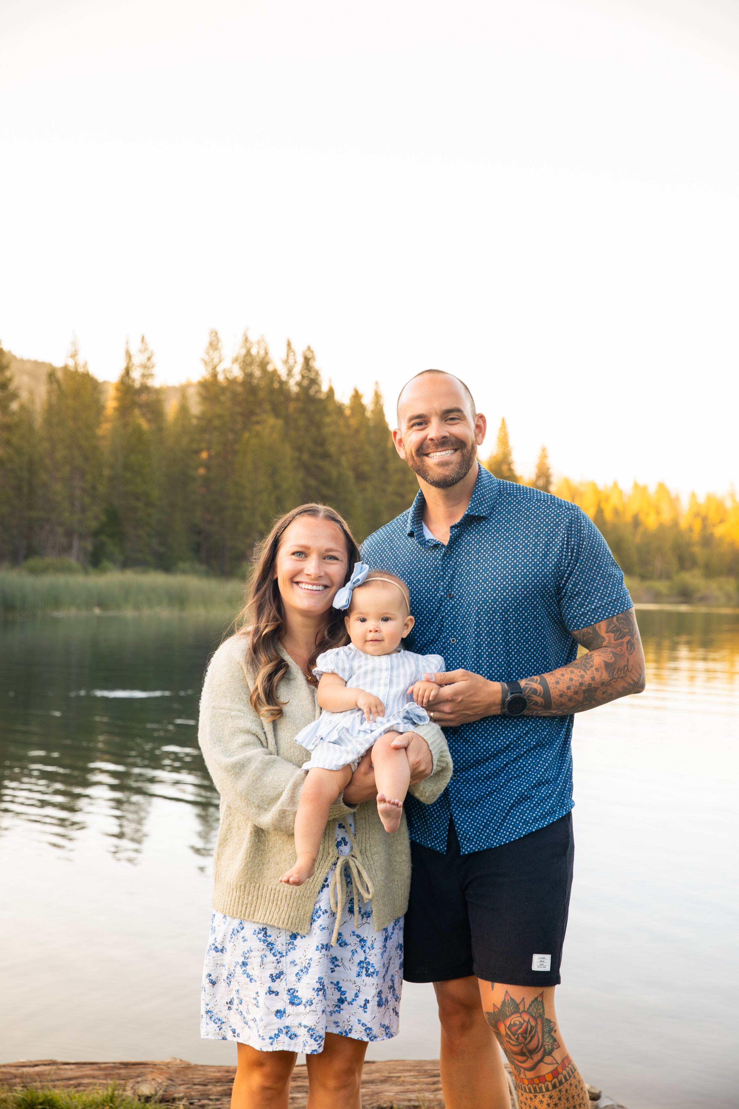 Family of three standing by a lake, smiling, with trees and a mountain in the background during sunset.