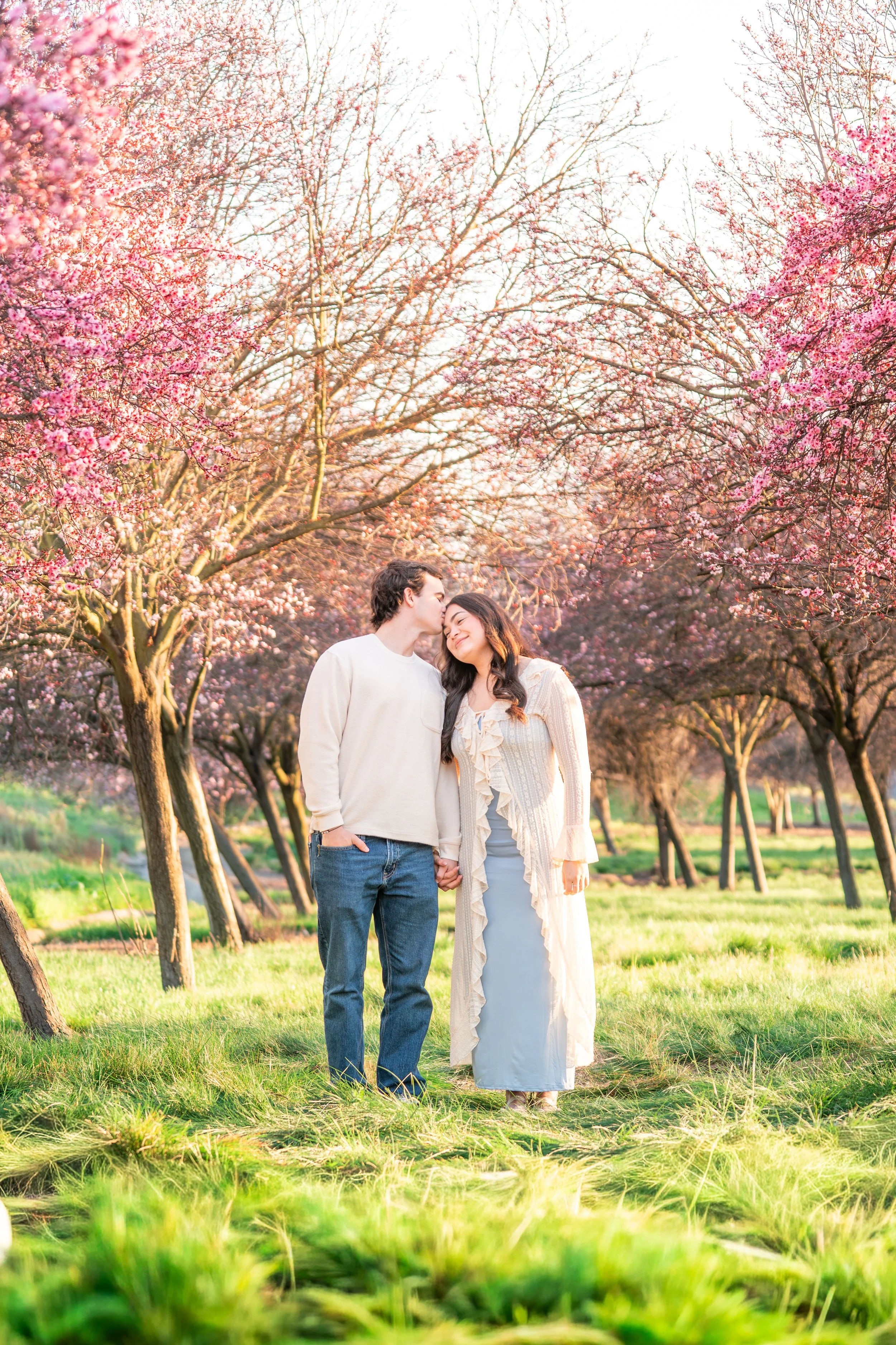 A couple holding hands in a park with pink blooming trees, bright green grass, and sunlight.