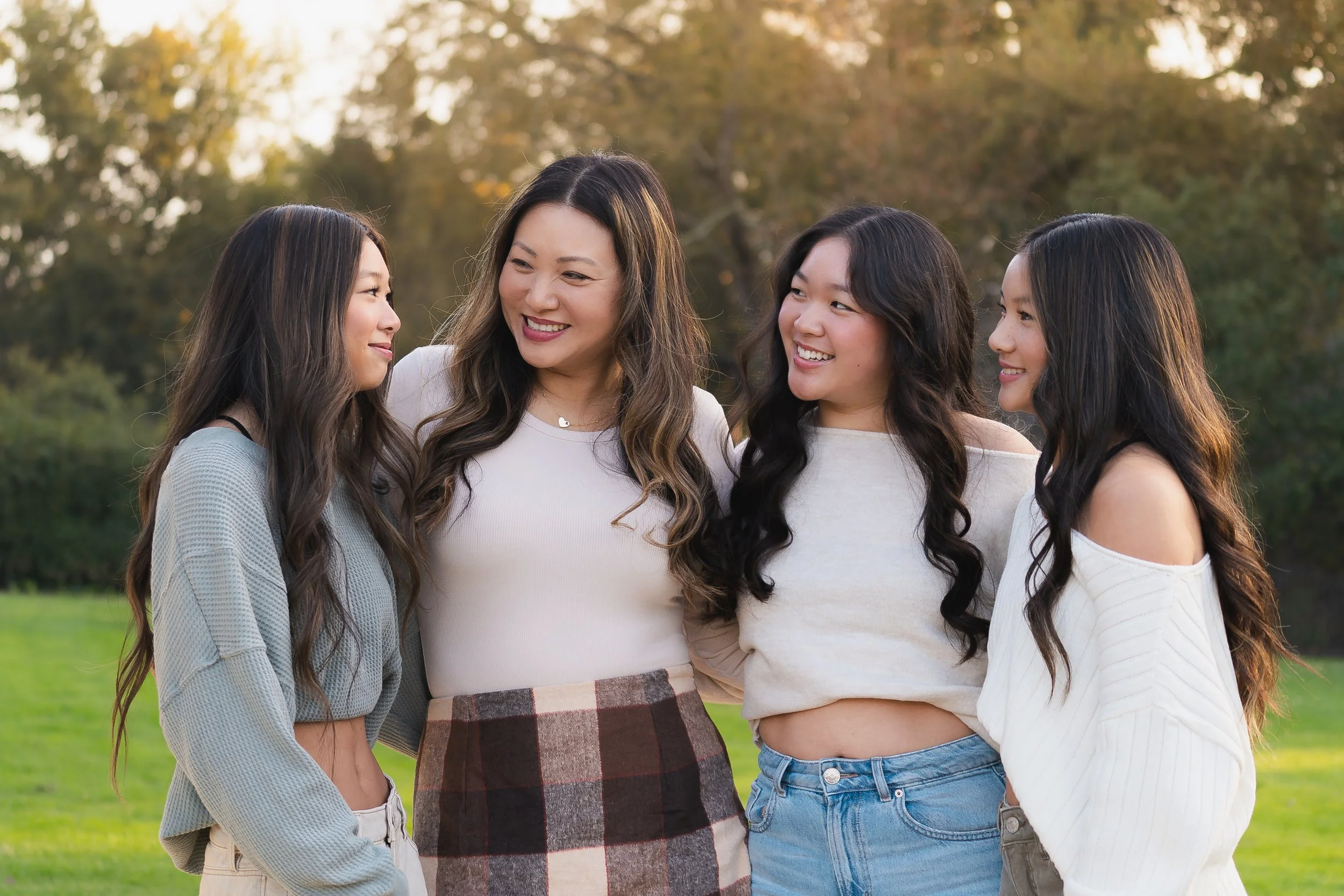A group of four women standing outdoors in a park, smiling and looking at each other, with trees and a green field in the background during autumn.