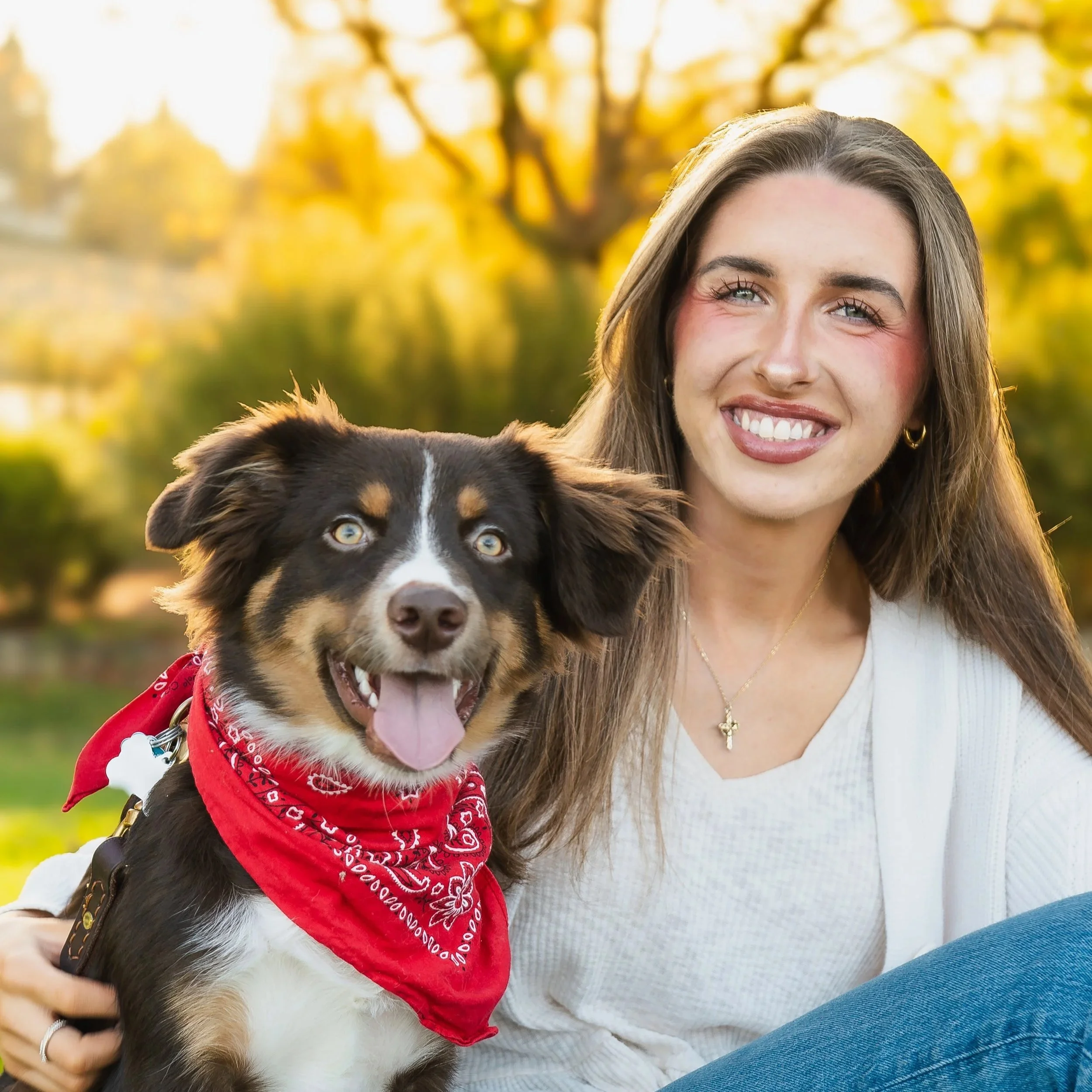 A smiling young woman with long brown hair and a dog in an outdoor park during fall, with trees having yellow leaves in the background.