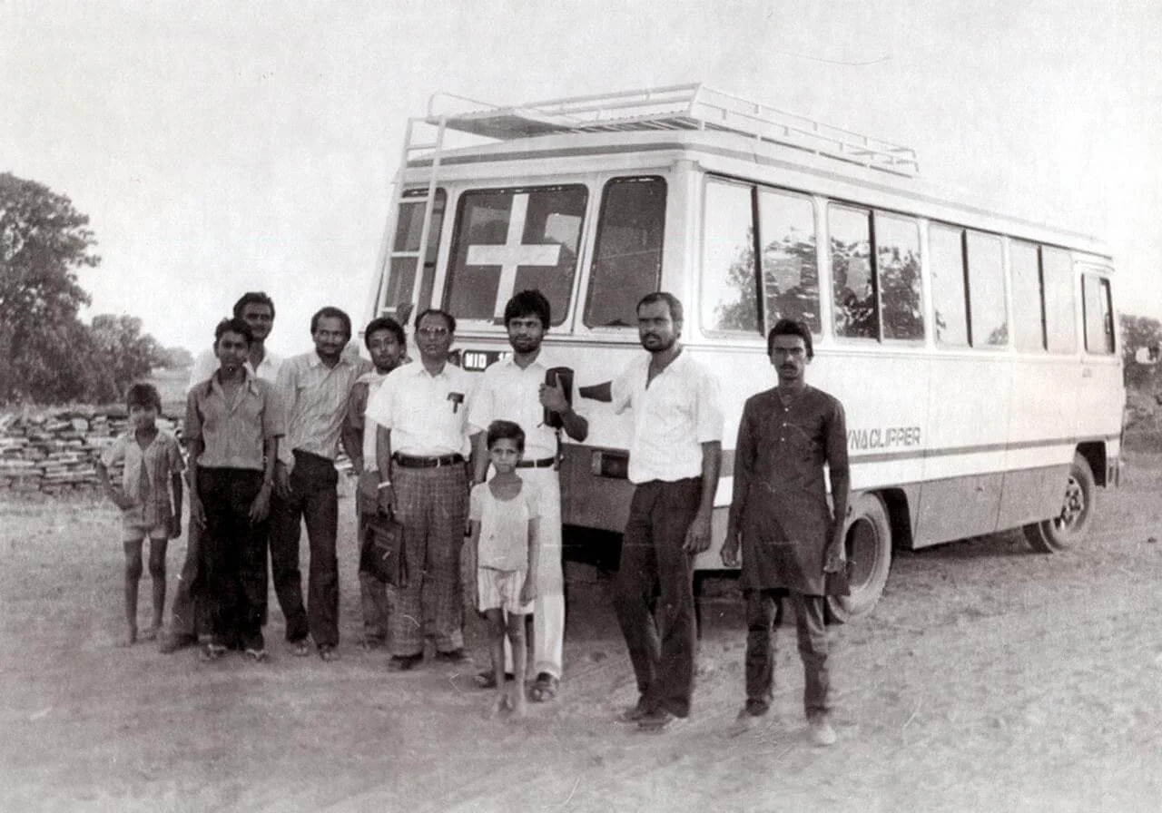 Group of men and children standing in front of an ambulance with a red cross symbol.