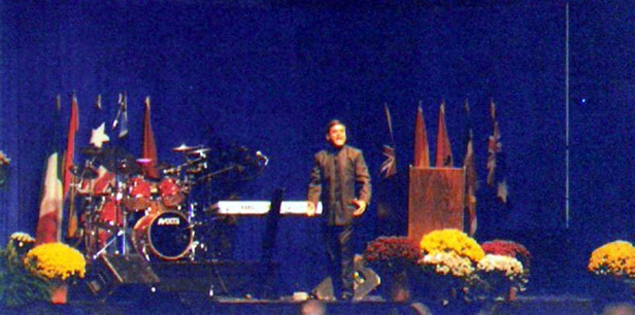 Man standing on stage at a formal event with musical instruments and flags in the background, surrounded by colorful flowers.